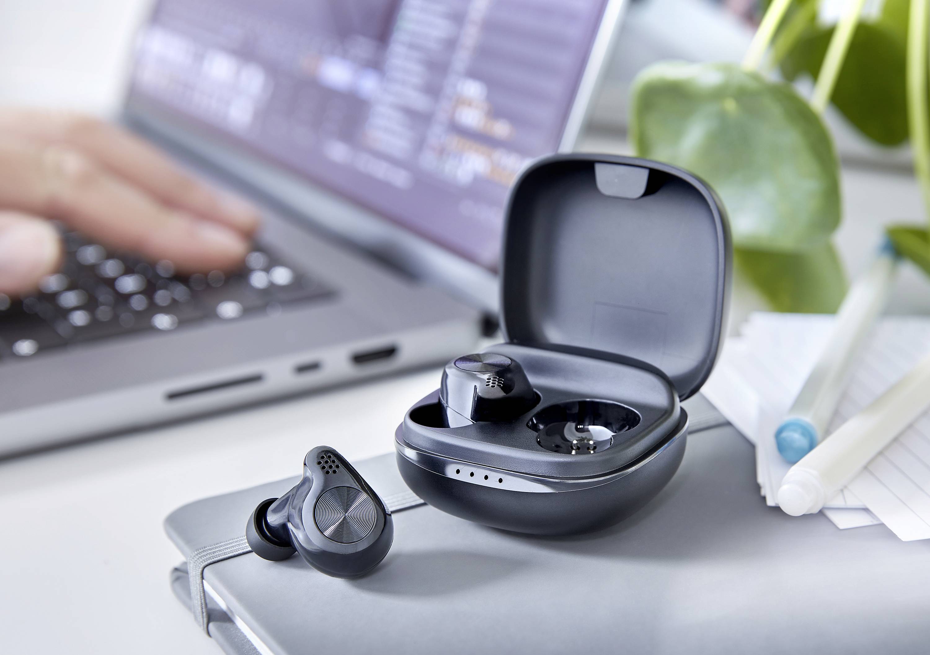 Draadloze oordopjes in een geopende oplaadcase staan op een laptop, terwijl een hand op de laptoptoetsenbord typt.