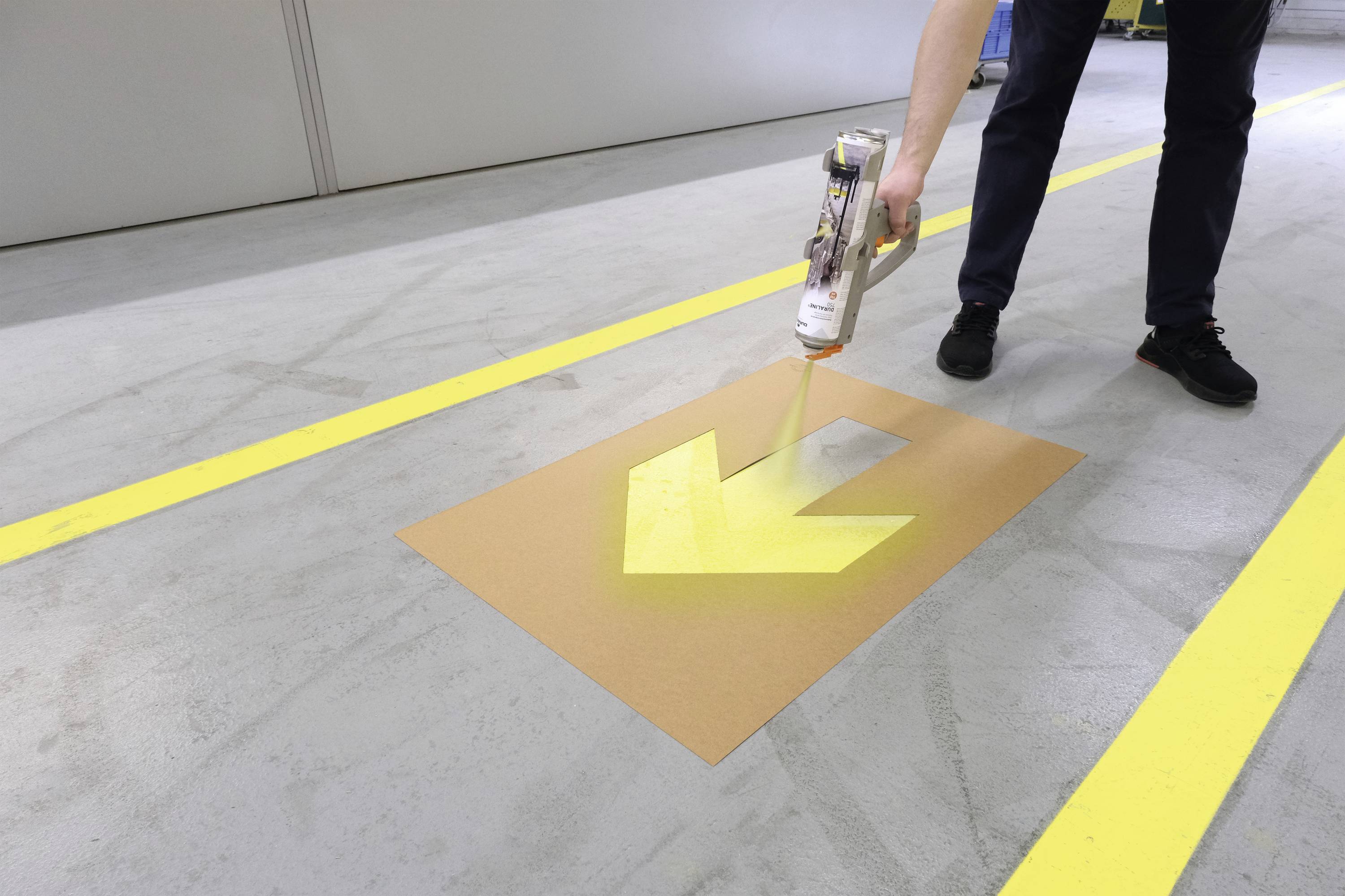 Een persoon spuit gele pijlen op de grond met behulp van een sjabloon om routes te markeren in een magazijn of industrieterrein.