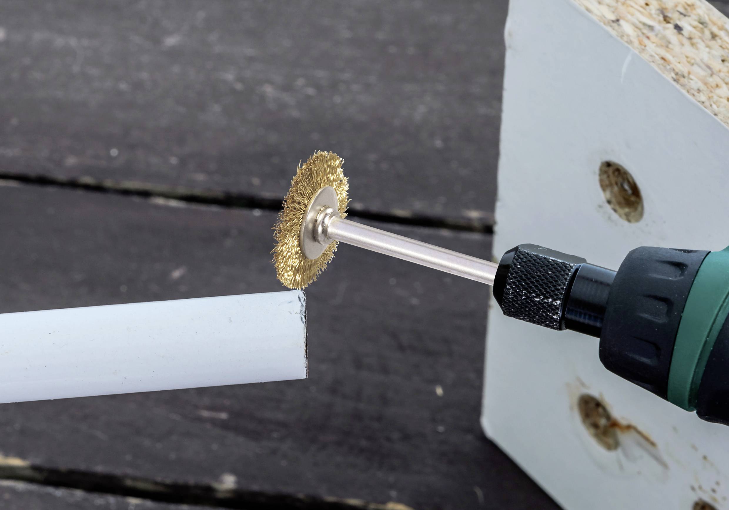 Een rotatietool met een draadborstelbijlage reinigt of polijst het uiteinde van een wit cilindrisch voorwerp, waarschijnlijk een pijp, nabij een houten blok.