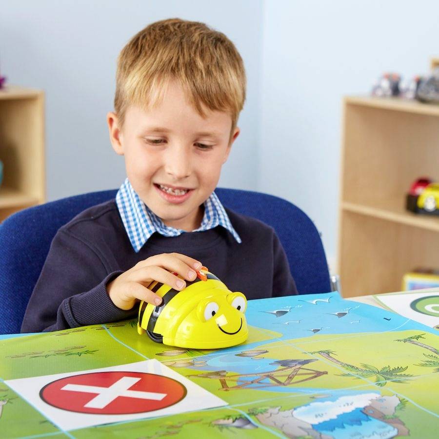 Een jongen speelt met een gele, bijenachtige robot op een leertapijt. Hij zit aan een tafel met leermaterialen.