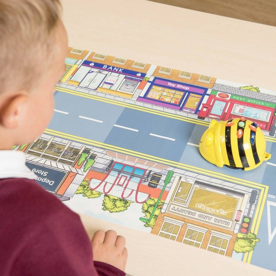 Een kind speelt met een gele, bijenachtige robot op een speelmat die een stadsstraat met verschillende winkels voorstelt.