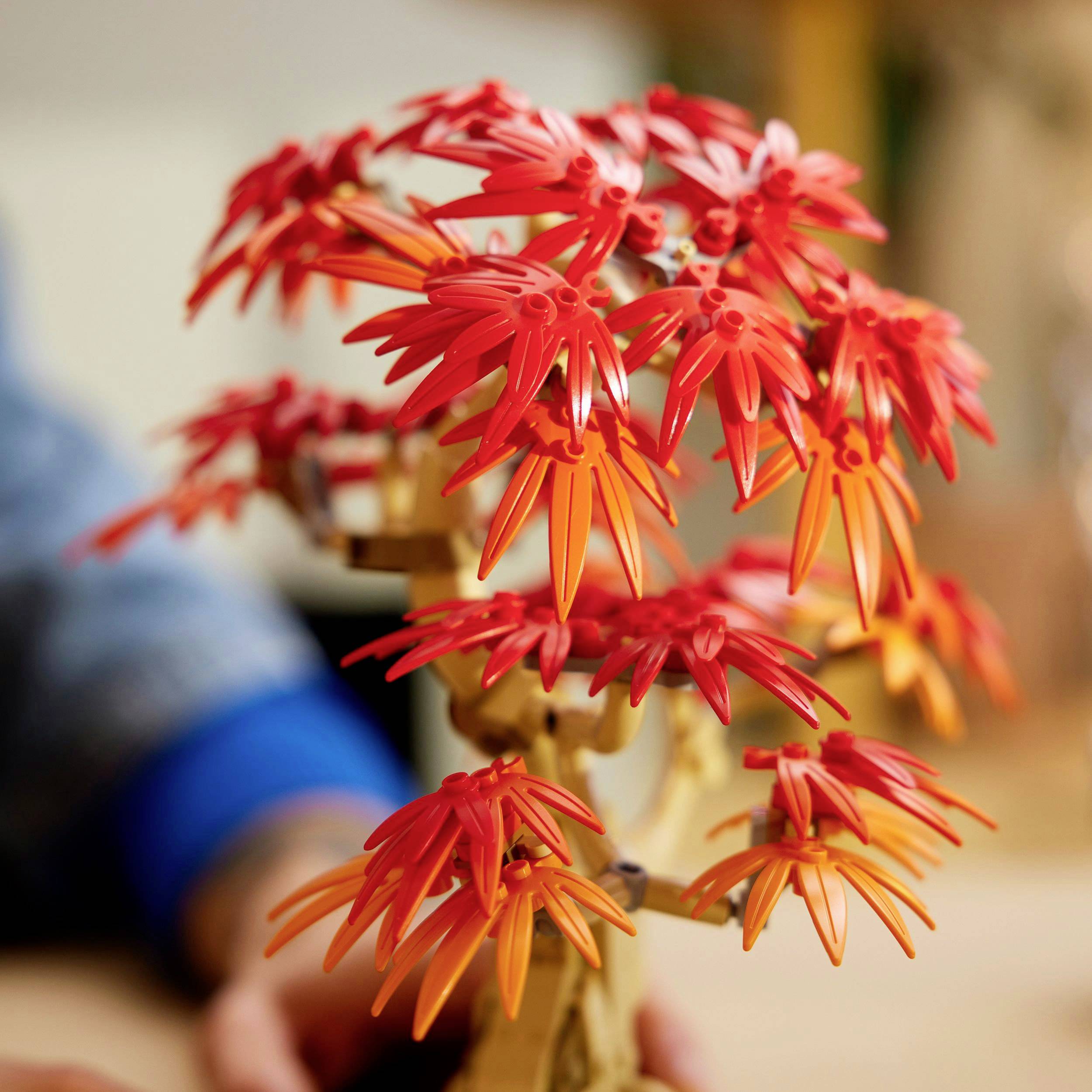 Een bonsai-boom van LEGO-stenen met felrode en oranje bladeren staat op een tafel. Handen op de achtergrond.