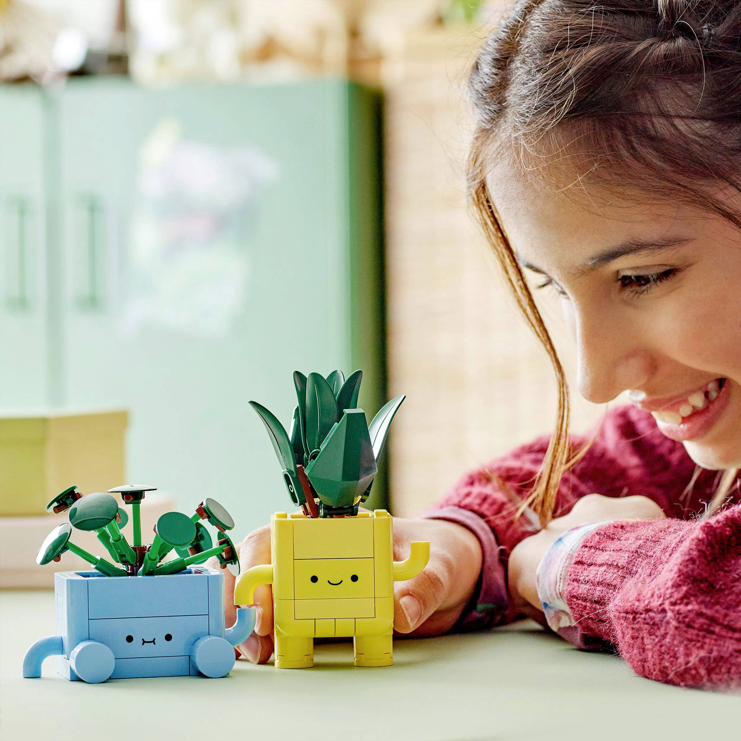 Een persoon zit glimlachend aan tafel en speelt met twee robot-plantenfiguren van Lego, een blauwe met bloemen en de andere geel met bladeren.