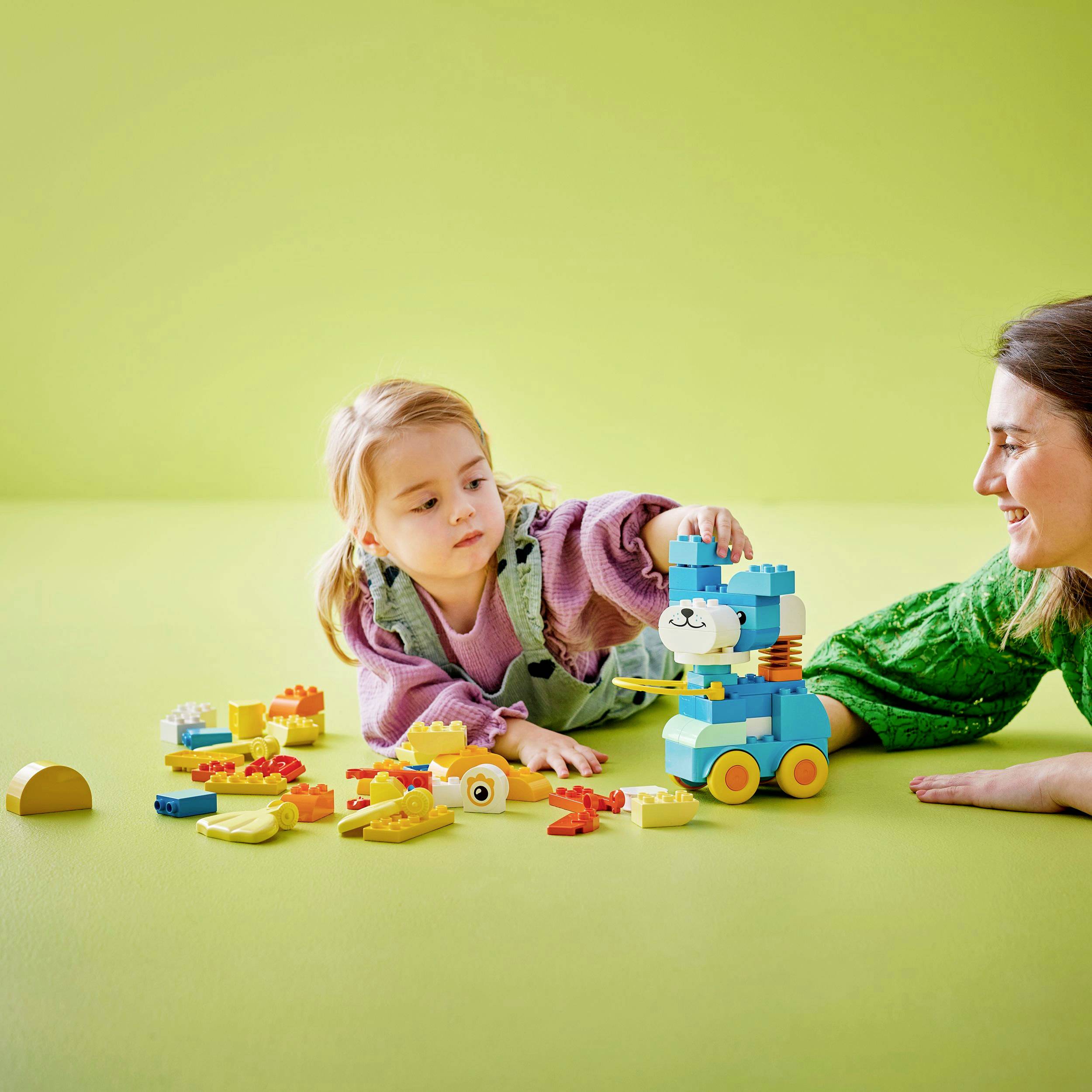 Een kind en een volwassene spelen met kleurrijke bouwblokken en een speelgoedauto op een gele tafel voor een groene achtergrond.