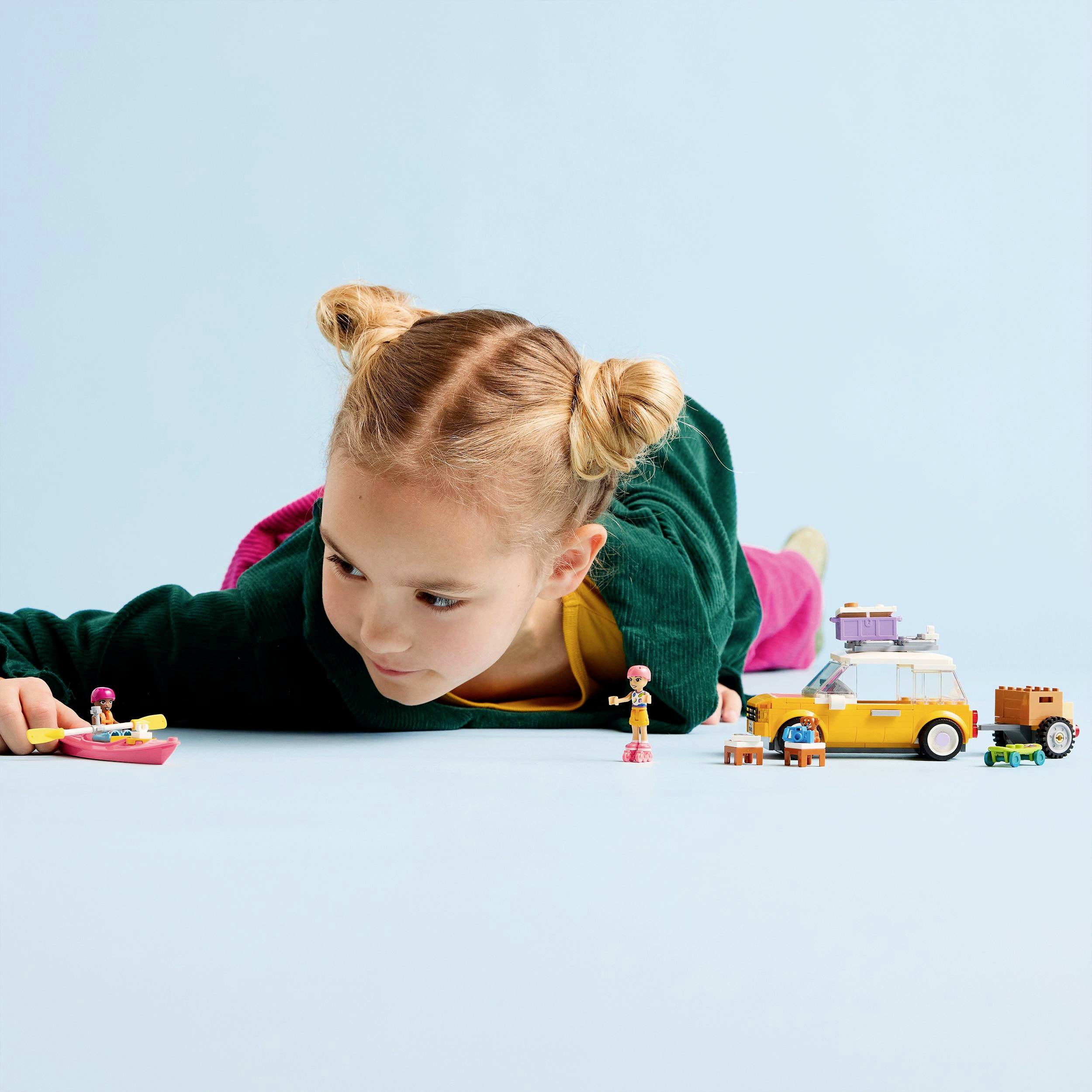 Een kind ligt op de grond en speelt met speelgoed, waaronder een gele speelgoedauto en figuren. Er heerst een speelse sfeer.