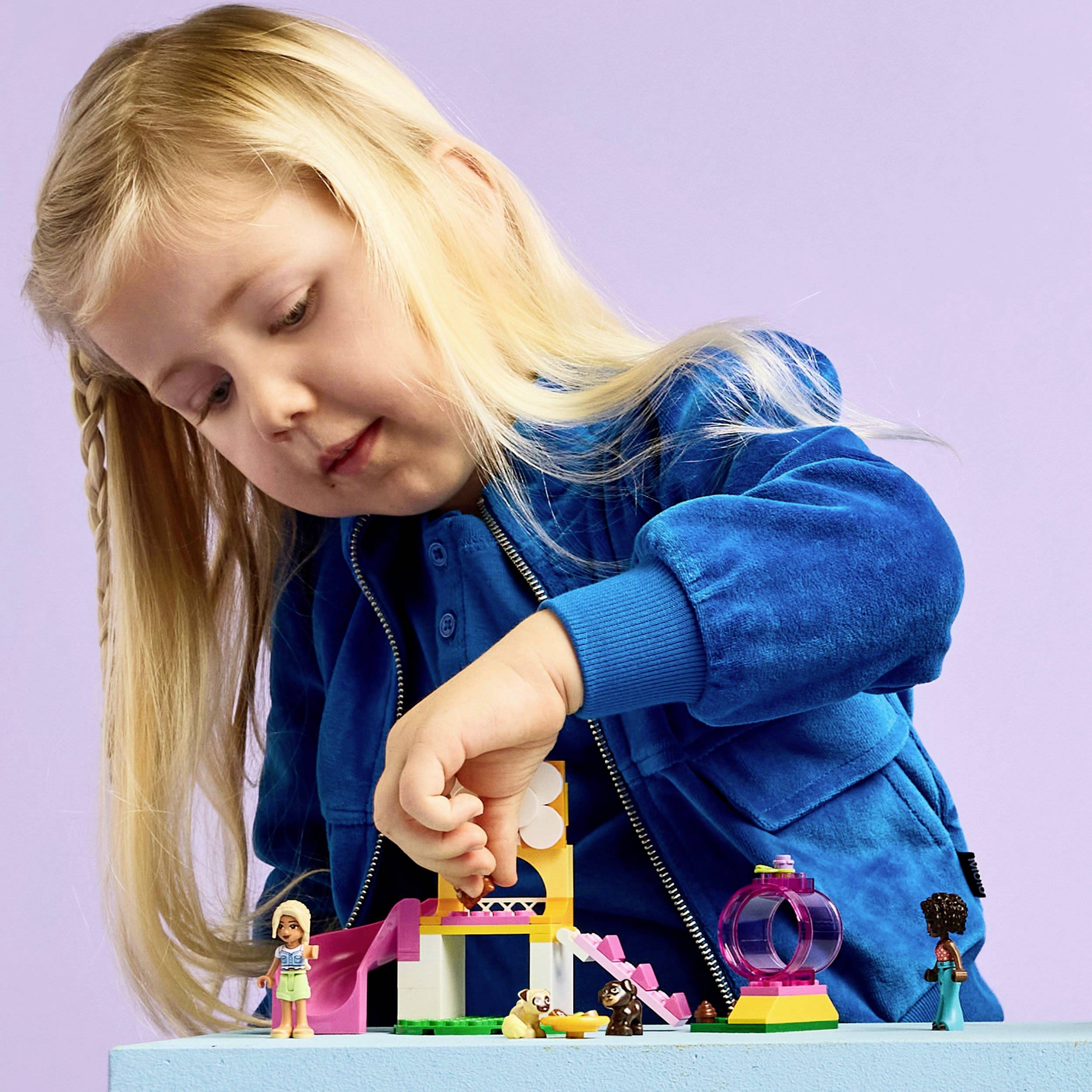 Een kind met lang blond haar speelt met kleurige bouwstenen en figuurtjes op een tafel. Het draagt een blauwe trui.