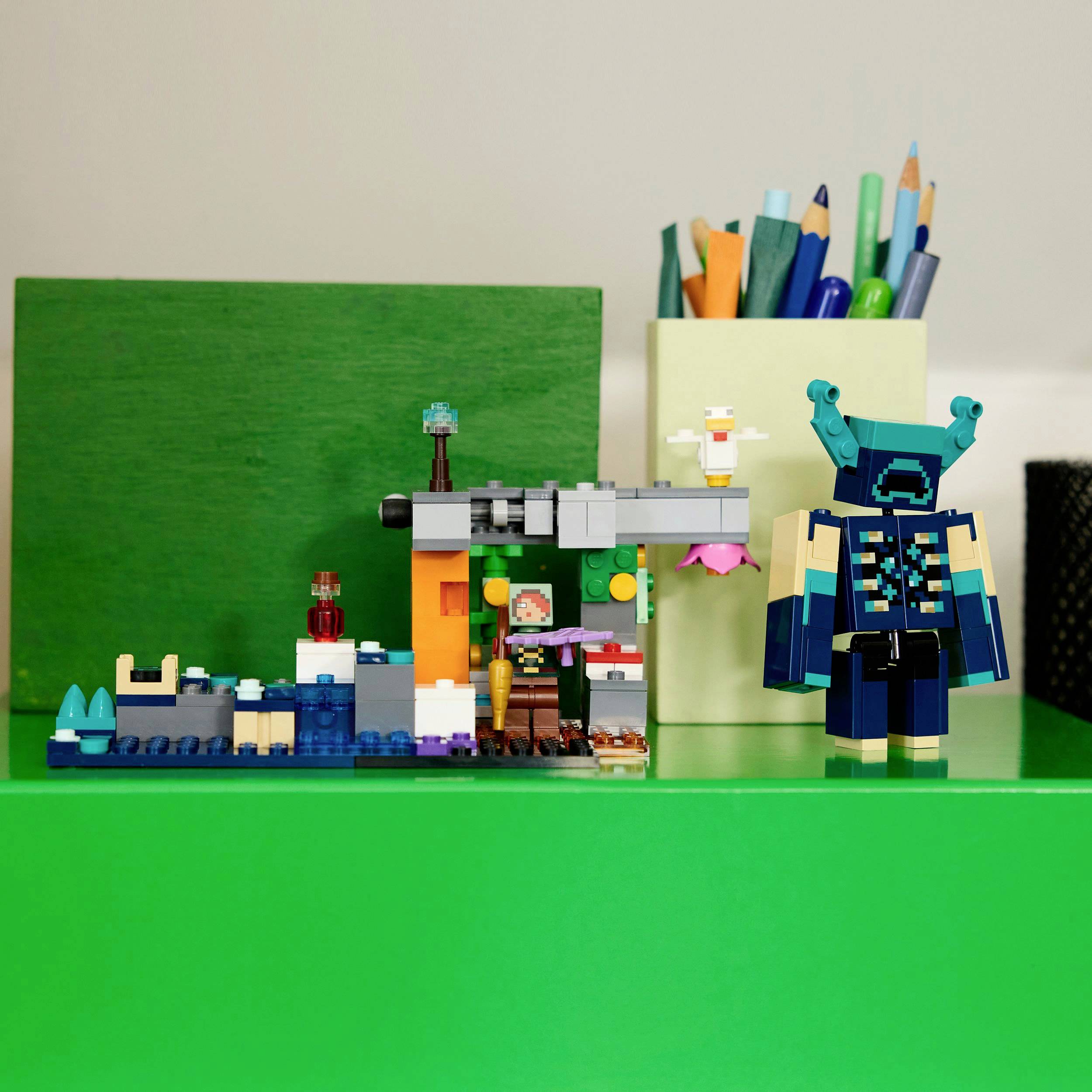 Kleurrijke Lego-modellen op een tafel. Links een scène met figuren en bouwwerken, rechts een groter, gedetailleerd model met pennen op de achtergrond.