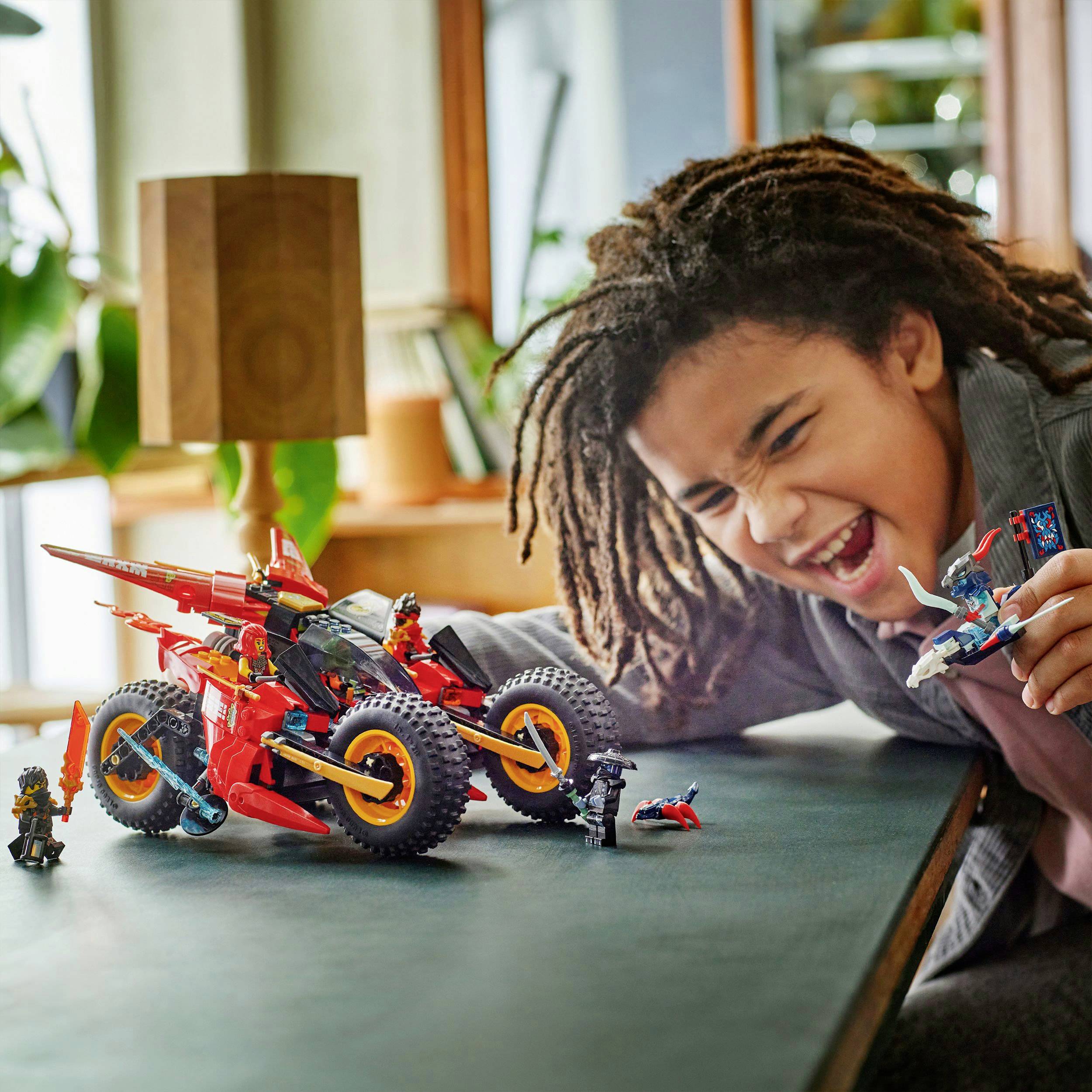 Een kind speelt enthousiast met kleurrijke speelgoedrobots op een tafel. Op de achtergrond zijn ramen en planten te zien.