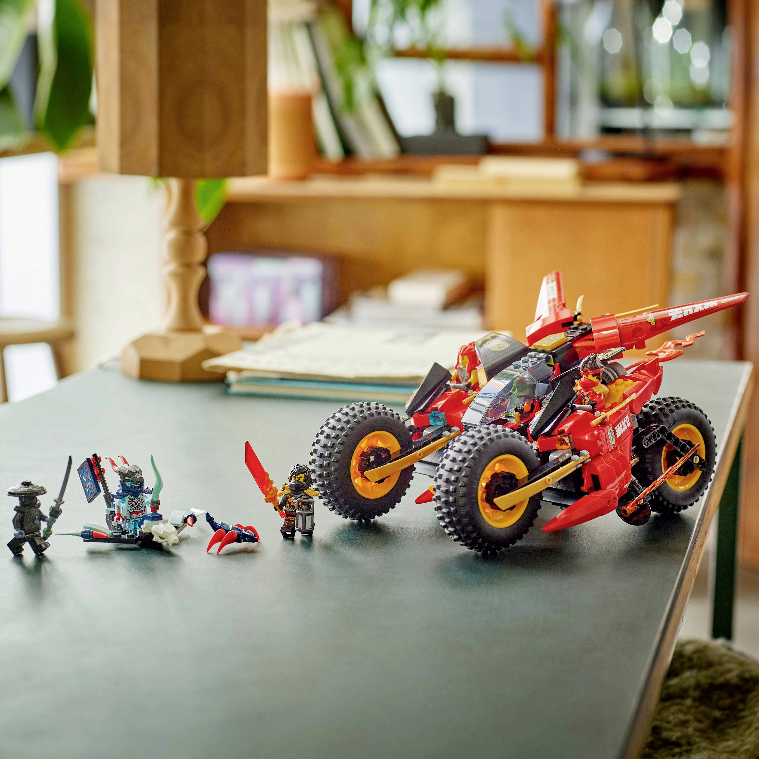 Drie kleurrijke speelgoed-actiefiguren en een modelvoertuig staan op een tafel in een kamer met planten en boeken op de achtergrond.