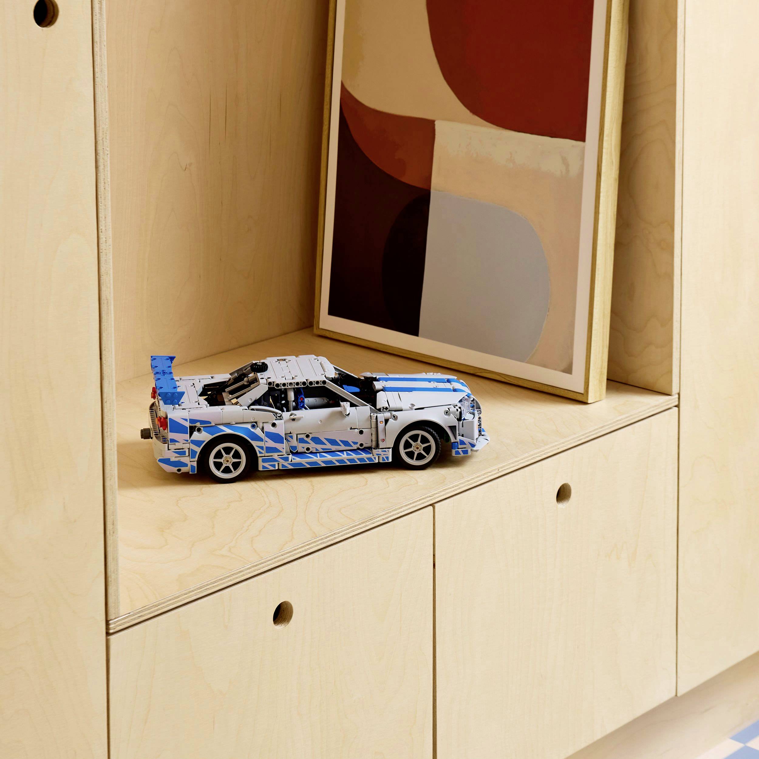 Een modelauto van blauwe kunststof onderdelen staat in een houten rek. Daarachter leunt een abstract schilderij tegen de rekwand.