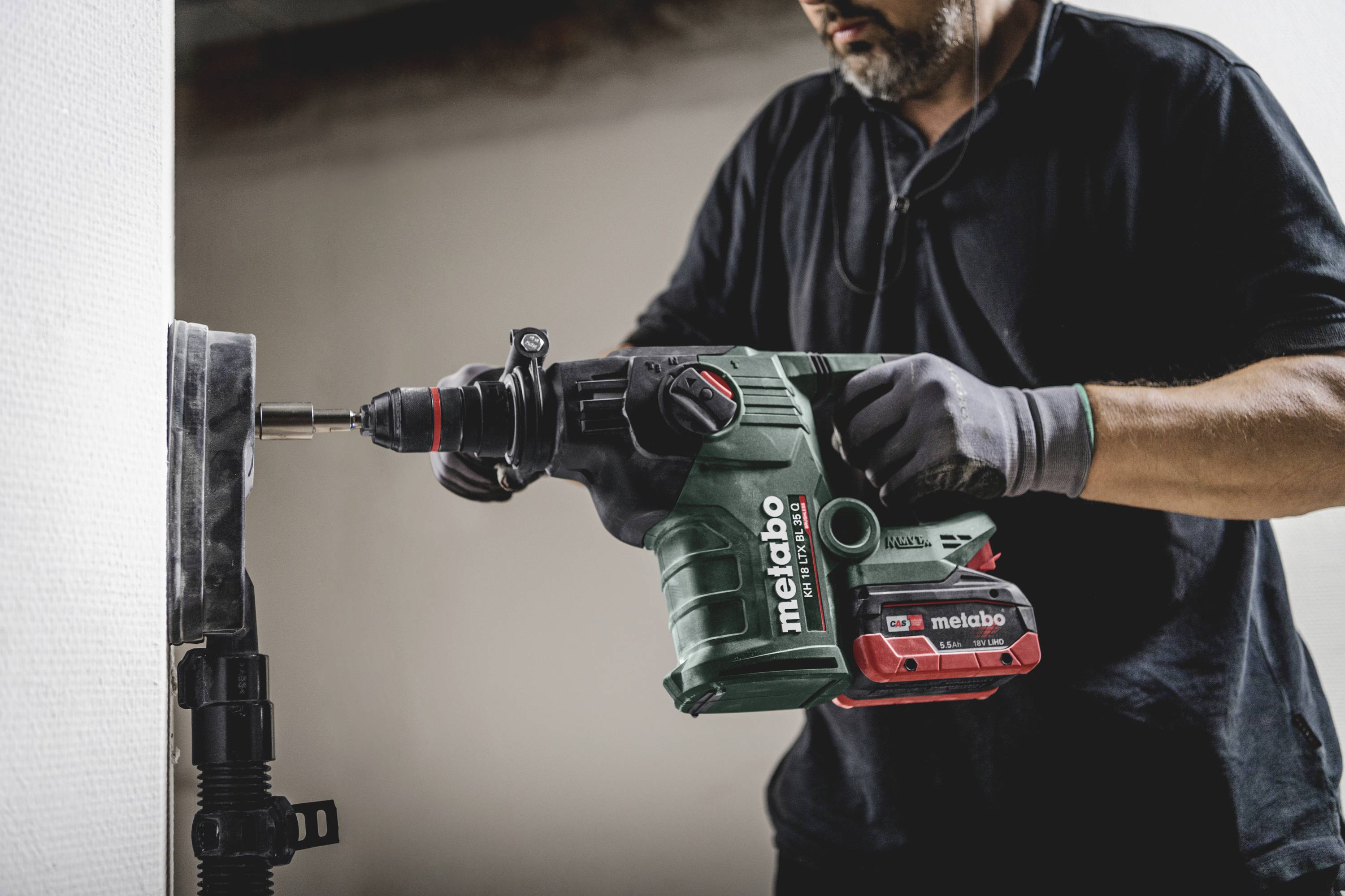 Een persoon boort met een groene Metabo-boorhamer een gat in de muur. De persoon draagt zwarte kleding en handschoenen.