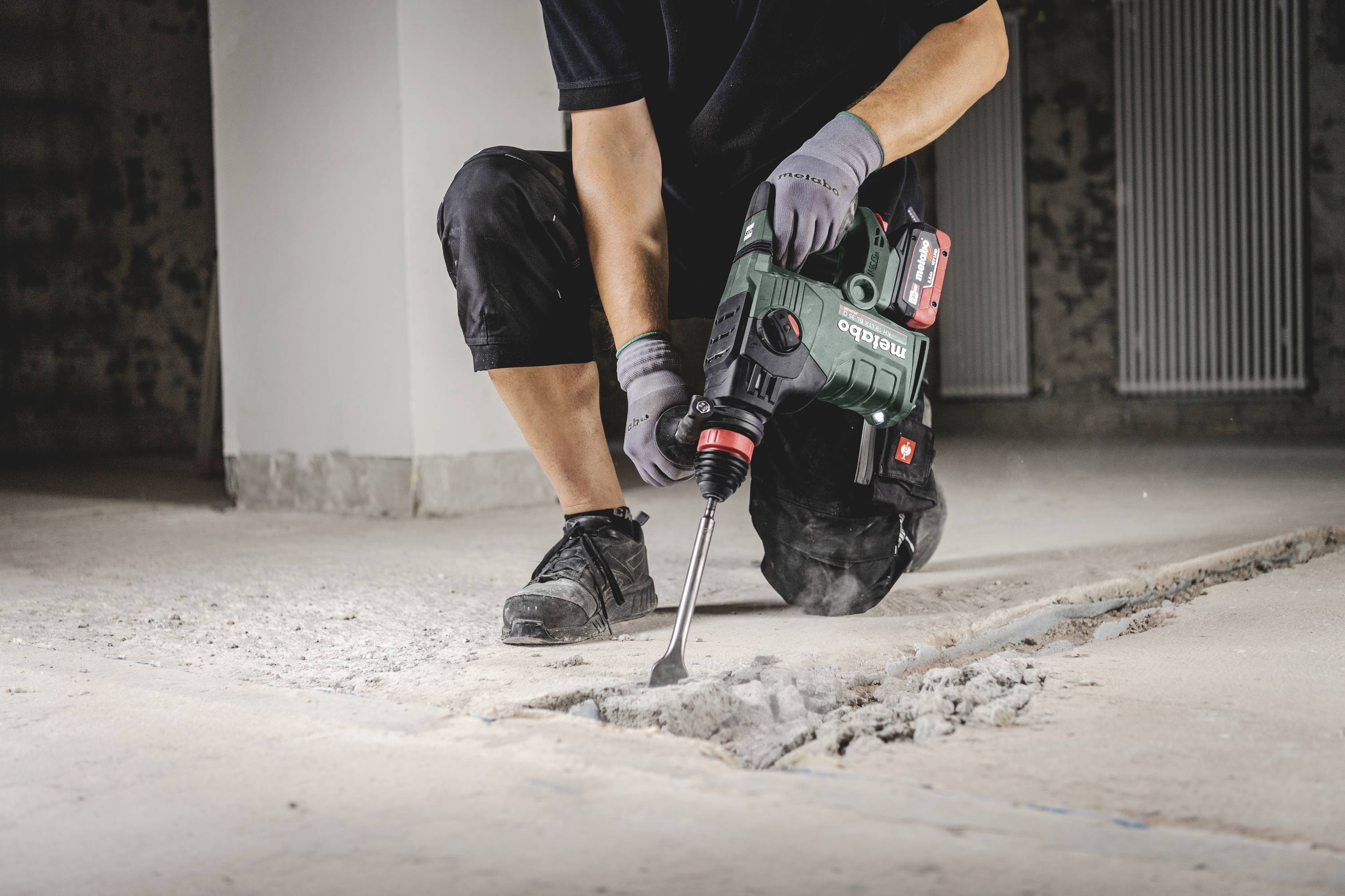 Een persoon gebruikt een groene boorhamer om betonvloer op te breken. De ruimte is onafgewerkt, met zichtbare muren en radiatoren.