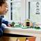 Een kind speelt met een LEGO-set aan een tafel bij een raam, met een huis, een boom en verschillende kleine poppetjes. Geschikt voor kinderen van 7 jaar en ouder.
