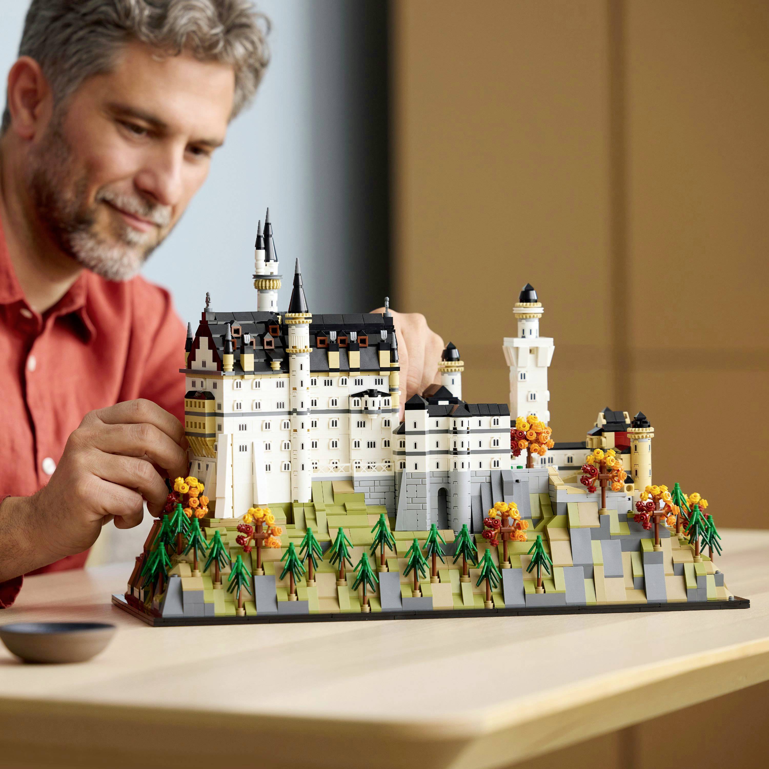 Een persoon bouwt een gedetailleerd LEGO-model van een kasteel aan een houten tafel, waarbij geïntreerde architectuur en levendige herfstige landschappen worden tentoongespreid.