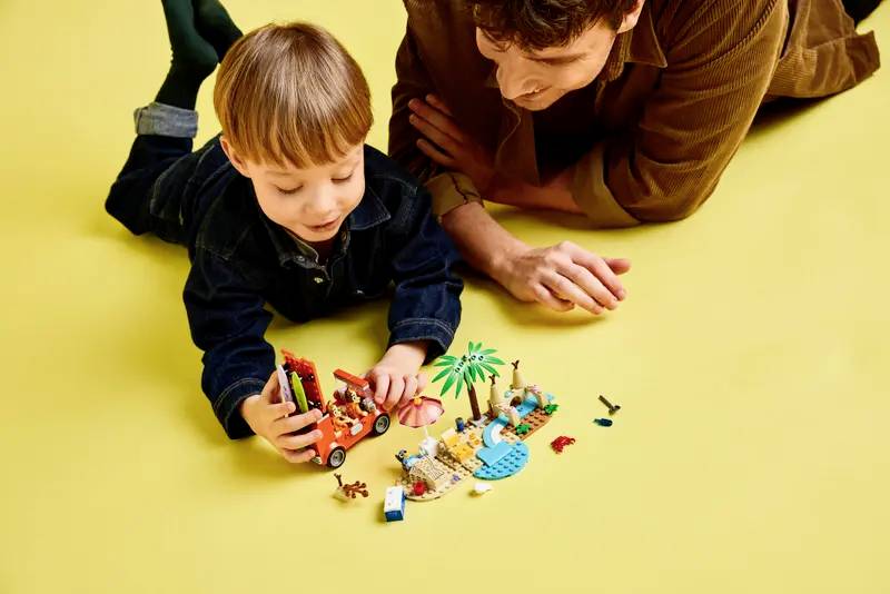 Een kind en een volwassene liggen op een gele vloer te spelen met kleurrijke blokken, waaronder een palmboom en kleine figuurtjes.