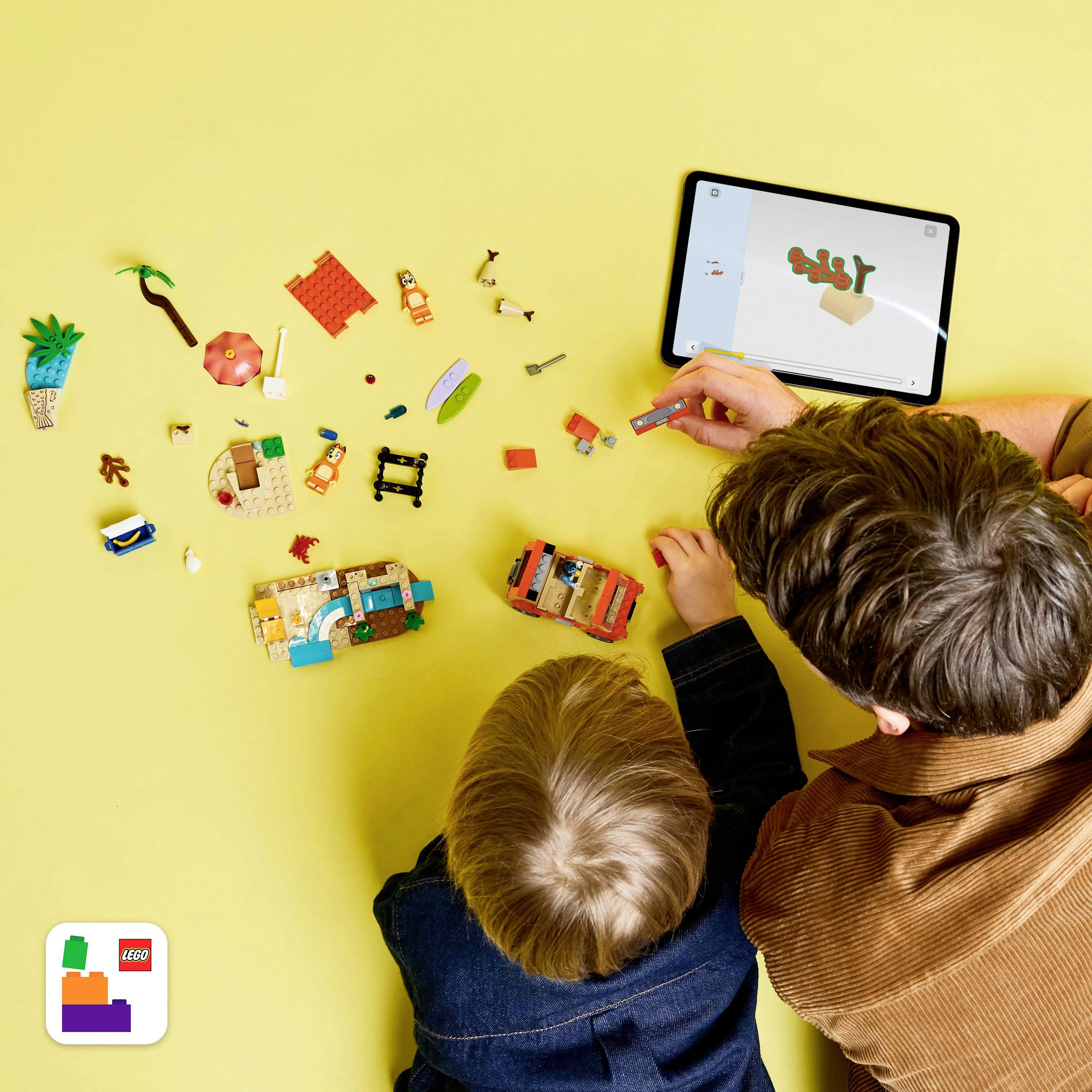 Een persoon en een kind bouwen Lego-stukken terwijl ze een tablet-app gebruiken die een 3D-model toont. Lego-stukken en miniaturen zijn verspreid over de tafel.