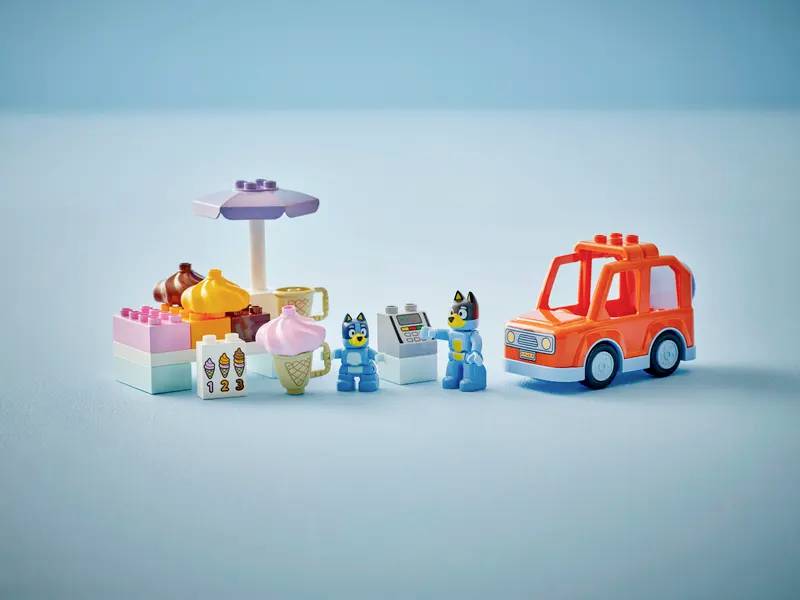 Speelgoedset met een kleine ijsjesstandaard, twee hondenfiguren en een rode speelgoedauto op een blauwe achtergrond, wat een speelse scène suggereert.