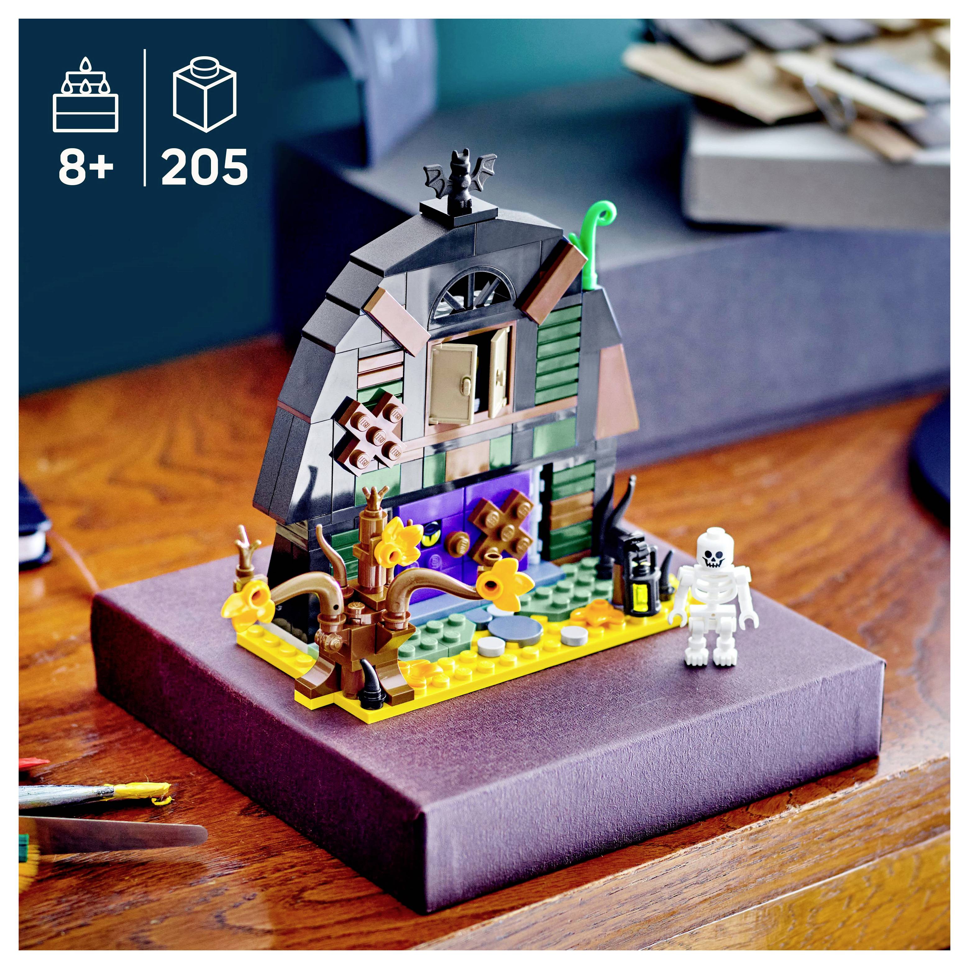 Een kleine, kleurrijke LEGO-spookhuis met verschillende details staat op een tafel. Een LEGO-skeletfiguur staat naast de set.