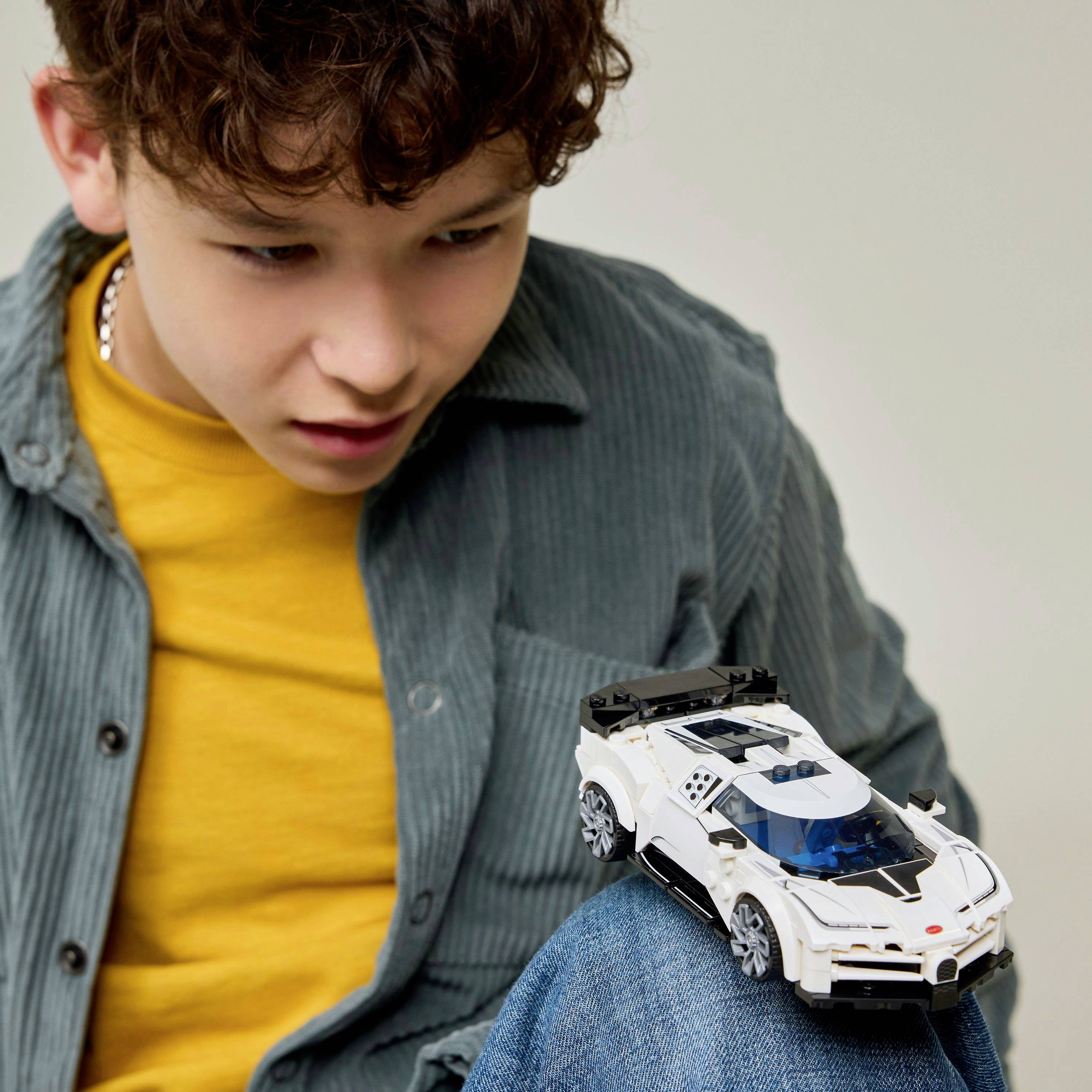 Een jongen in een geel shirt en een grijze jas kijkt nieuwsgierig naar een witte speelgoedauto die op zijn knie staat. De focus is gericht op de speelgoedauto.