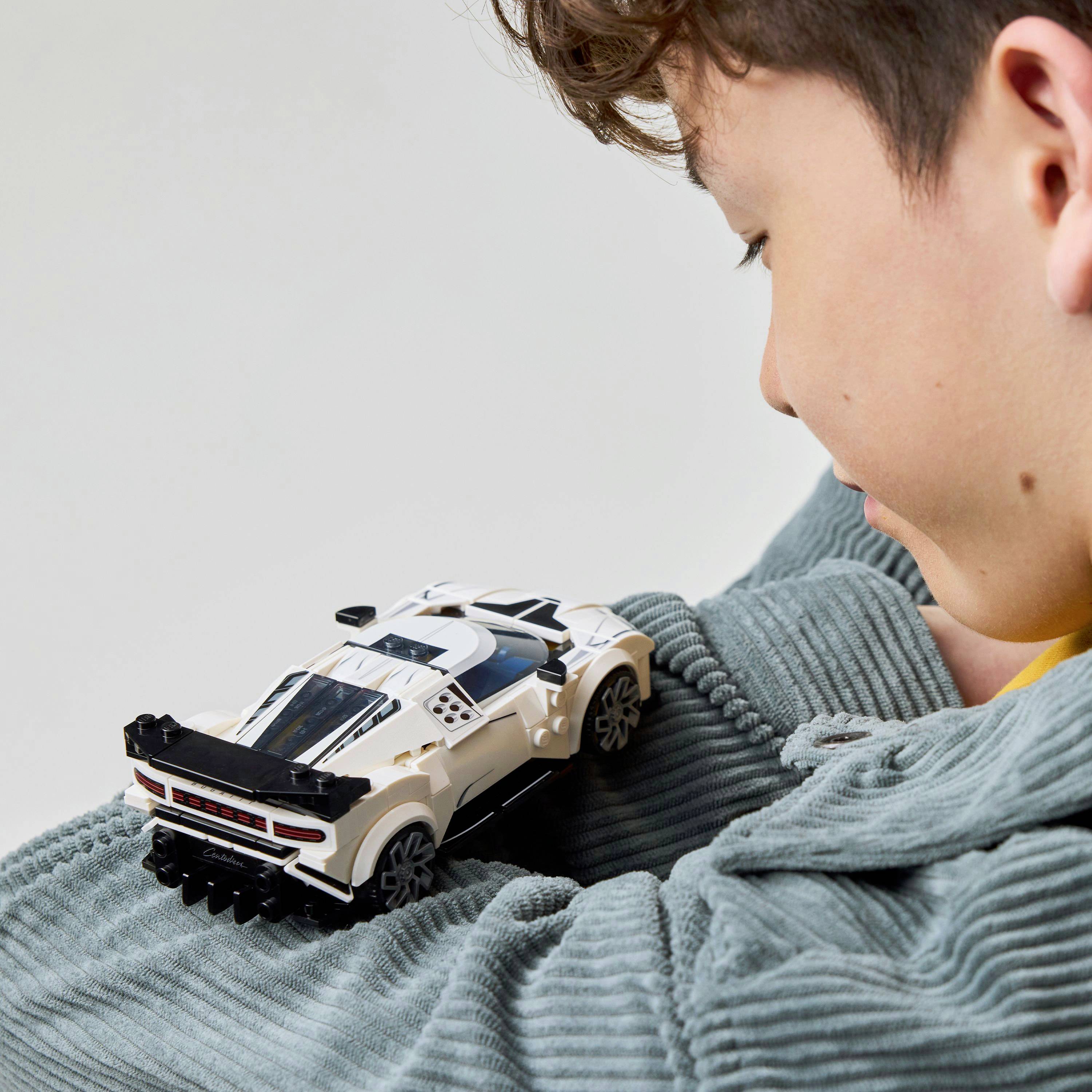 Een kind dat met een witte speelgoedauto op zijn arm speelt. Het kind glimlacht licht en draagt een gestructureerde, grijze trui.