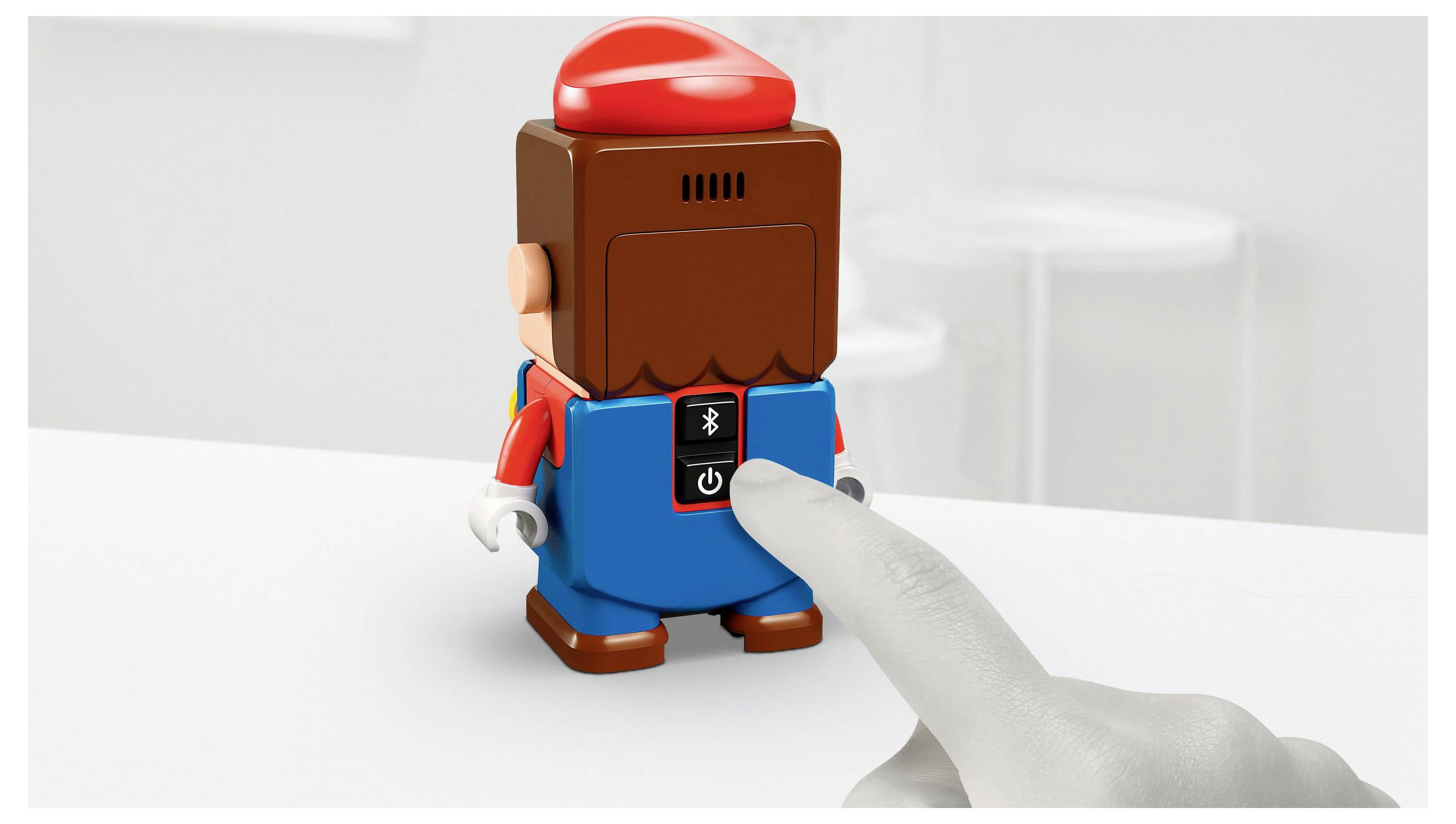 Een hand drukt op de knop aan de achterkant van een speelfiguur gekleed in blauw en rood, waarschijnlijk om hem te activeren. Het speelgoed staat op een wit oppervlak.