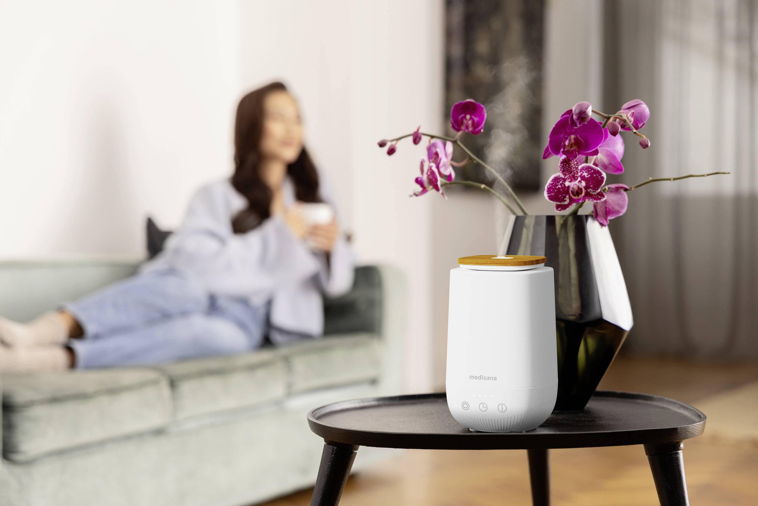 Een luchtreiniger staat op een tafel naast een vaas met paarse orchideeën. Op de achtergrond zit een onscherpe persoon op een bank.