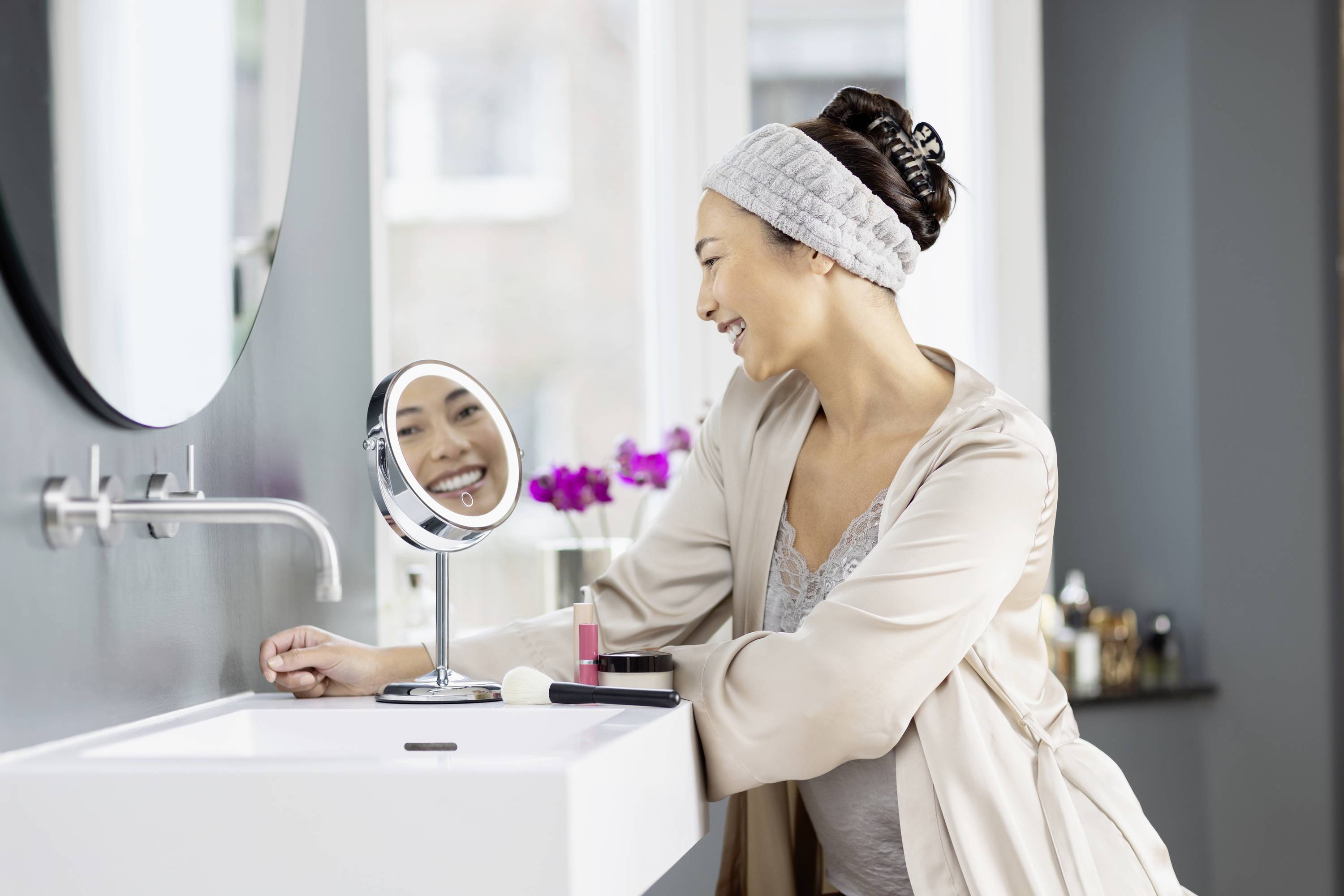 Vrouw glimlacht in een modern badkamerspiegeltje. Ze draagt een badjas en een haarband. Cosmeticaproducten liggen op de wastafel.