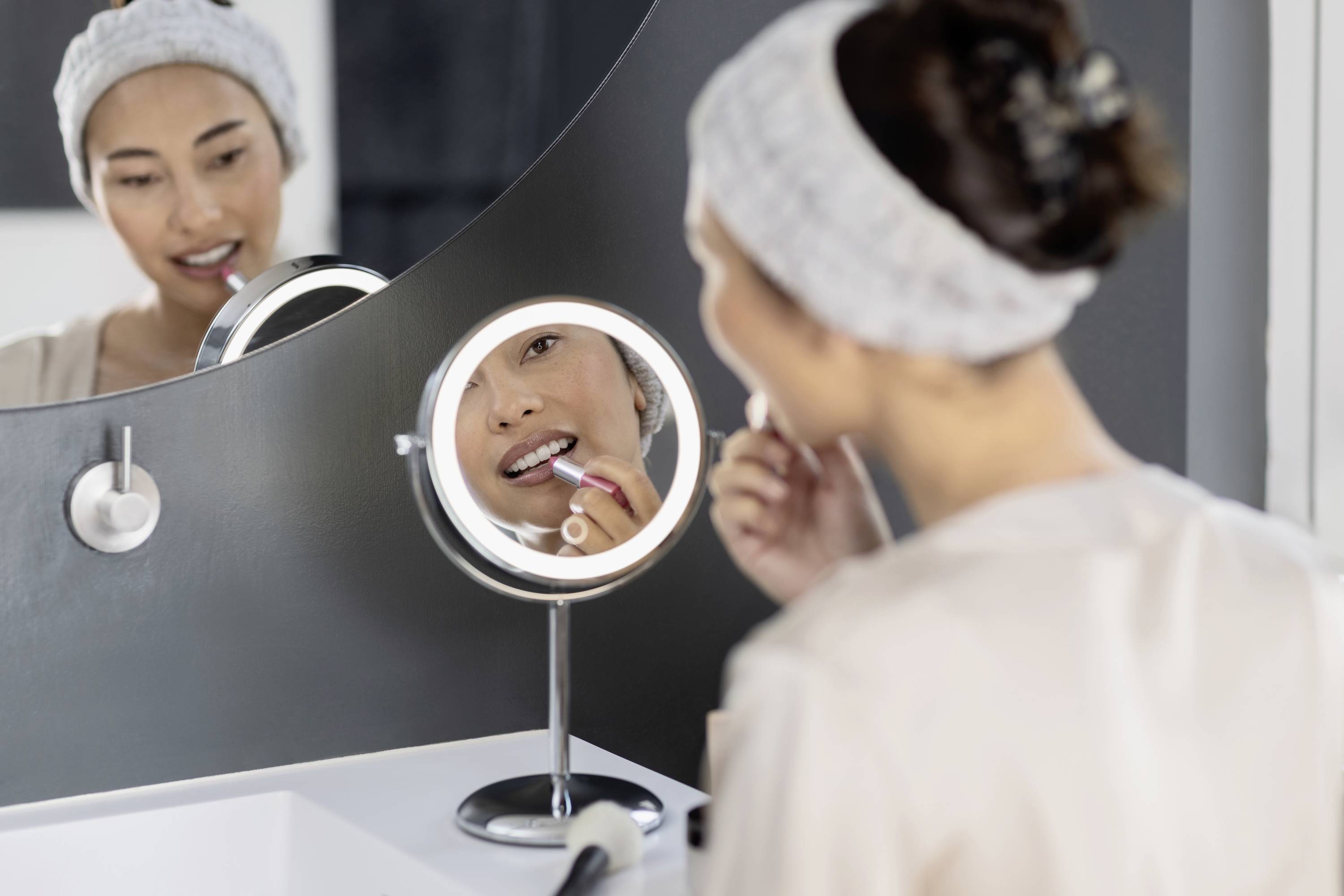 Een vrouw brengt lippenstift aan voor een ronde spiegel in de badkamer. Ze draagt een haarband en kijkt in de spiegel.