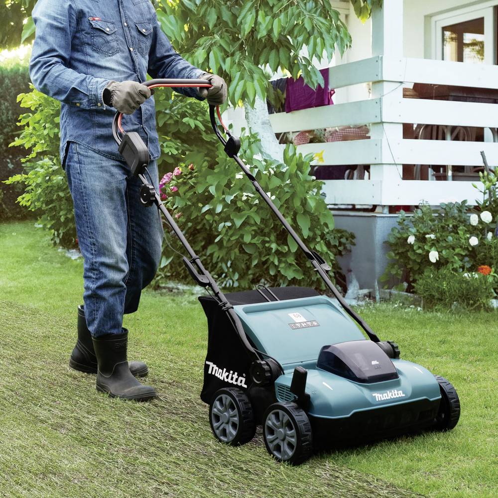 Een persoon maait het gazon met een elektrische grasmaaier op een groene weide in een tuin. Op de achtergrond zijn struiken en een huis te zien.