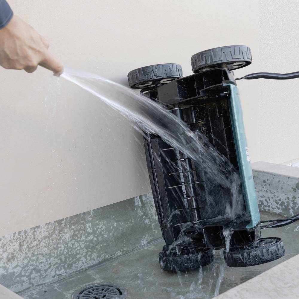 Een stofzuigerrobot wordt schoongemaakt met water terwijl hij ondersteboven ligt. Een hand houdt de waterslang vast.