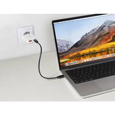 Een laptop op een tafel wordt opgeladen, aangesloten op een stopcontact. Het laptopscherm toont een berglandschap als achtergrond en verschillende app-pictogrammen.