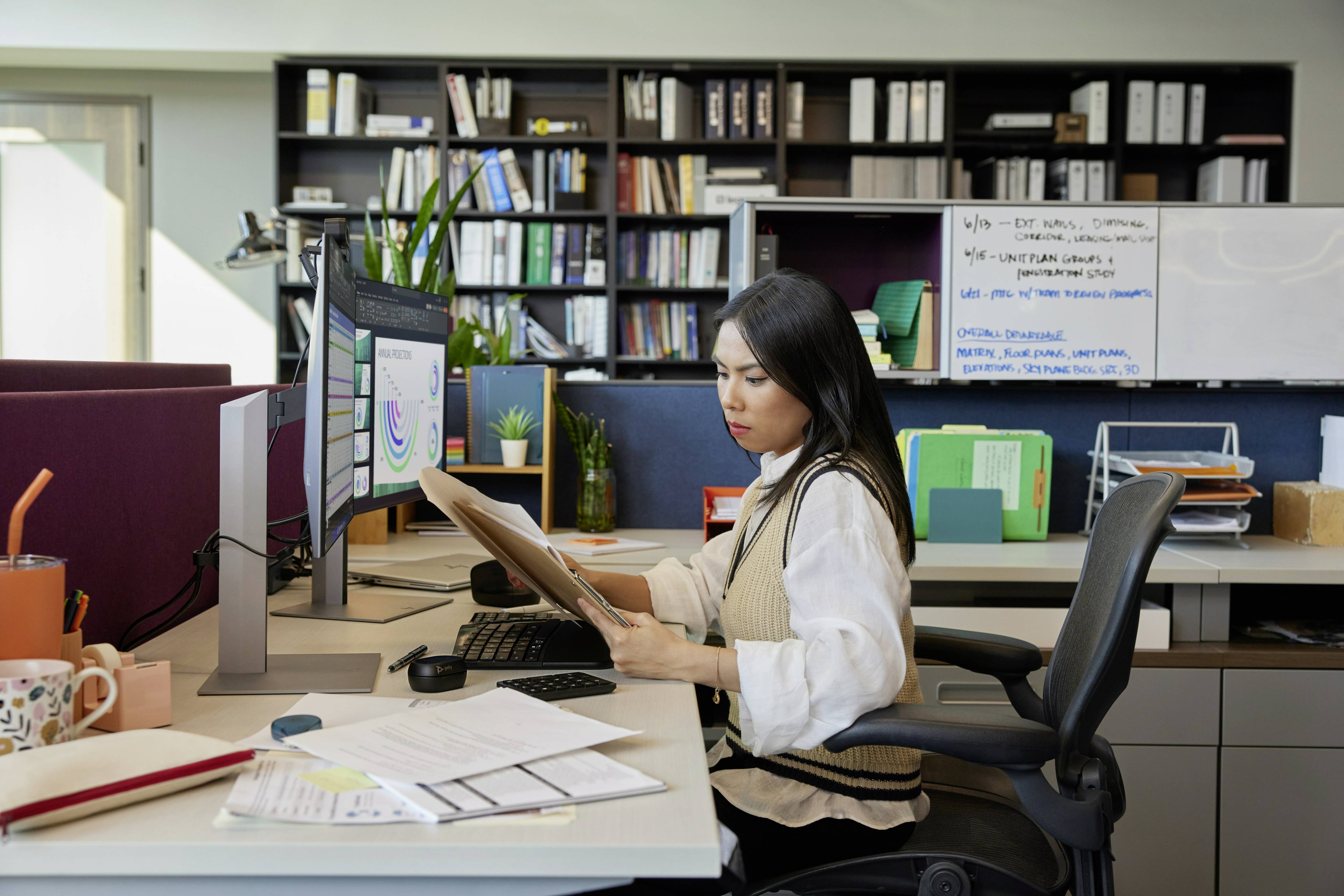 Een vrouw zit aan een bureau in een kantoor, houdt een klembord vast en kijkt naar een computerscherm. Op de achtergrond zijn boekenkasten met mappen.
