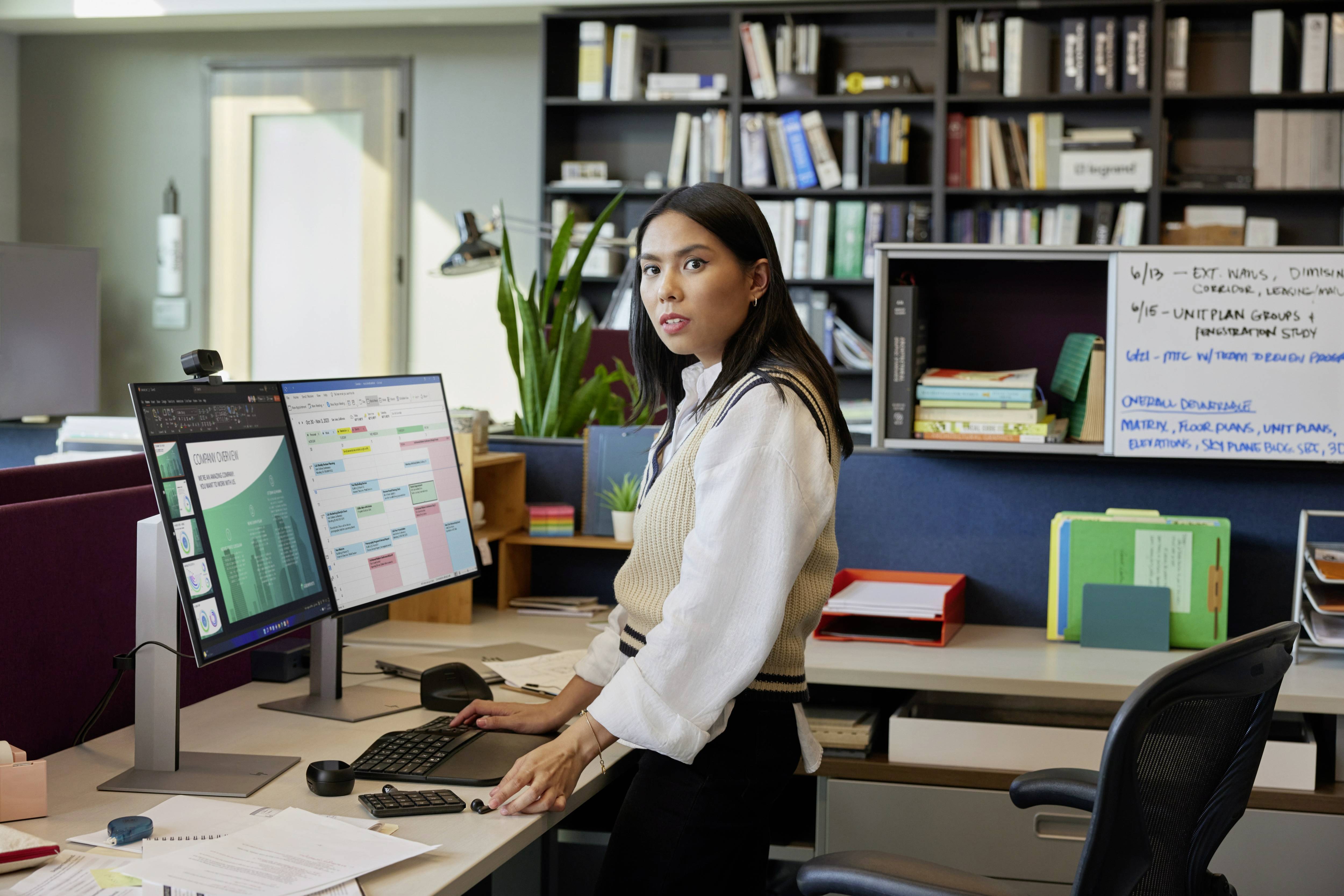 Een vrouw staat bij een bureau in een kantoor, kijkt naar twee computerschermen en is omgeven door planken en notities.