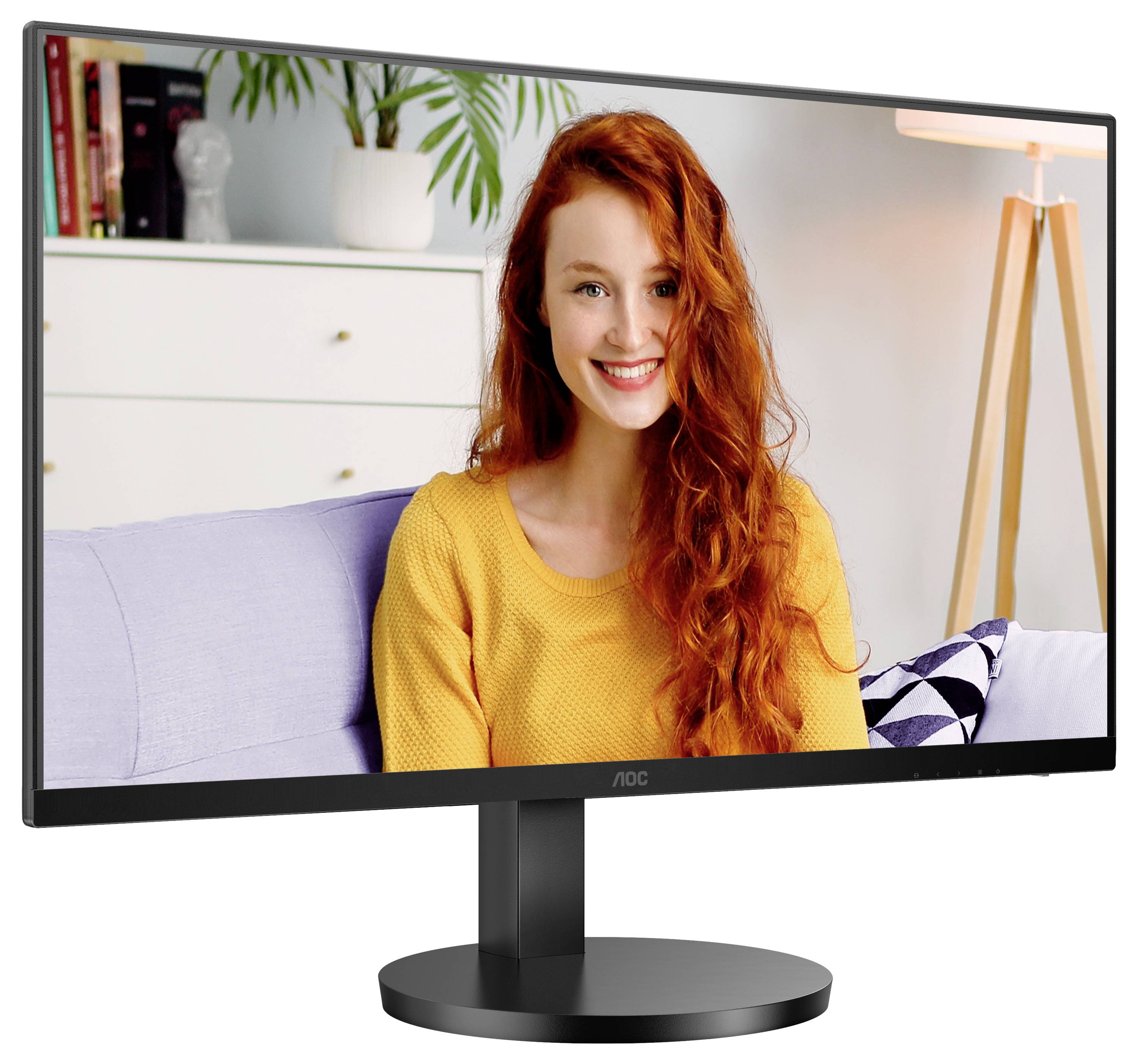 Een vrouw met rood haar en een gele trui glimlacht op een computermonitor in een woonkamer met een lamp en een ladenkast op de achtergrond.
