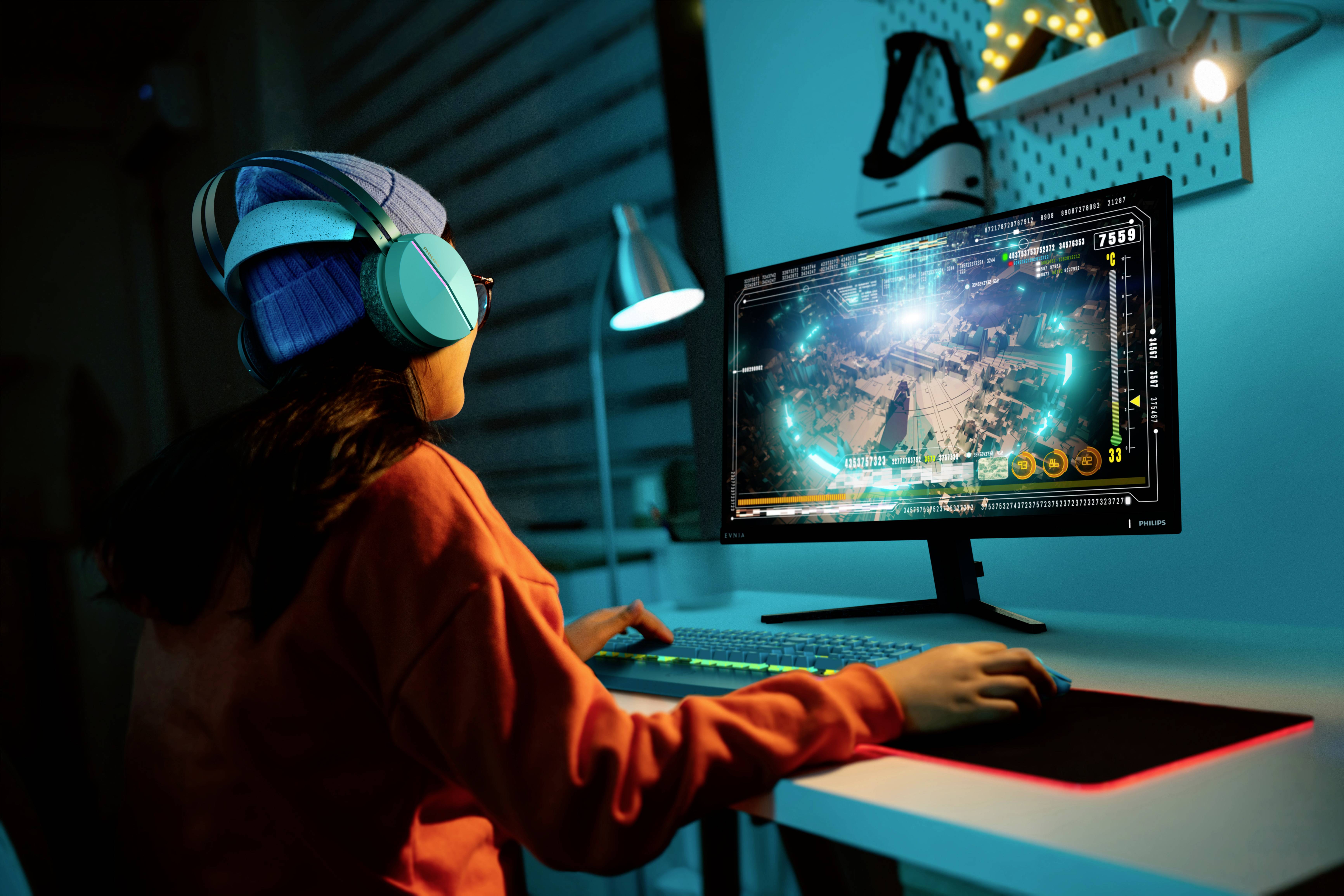 Een persoon met koptelefoon en muts speelt op een computer. Het scherm toont een strategiespel met veel details en getallen.