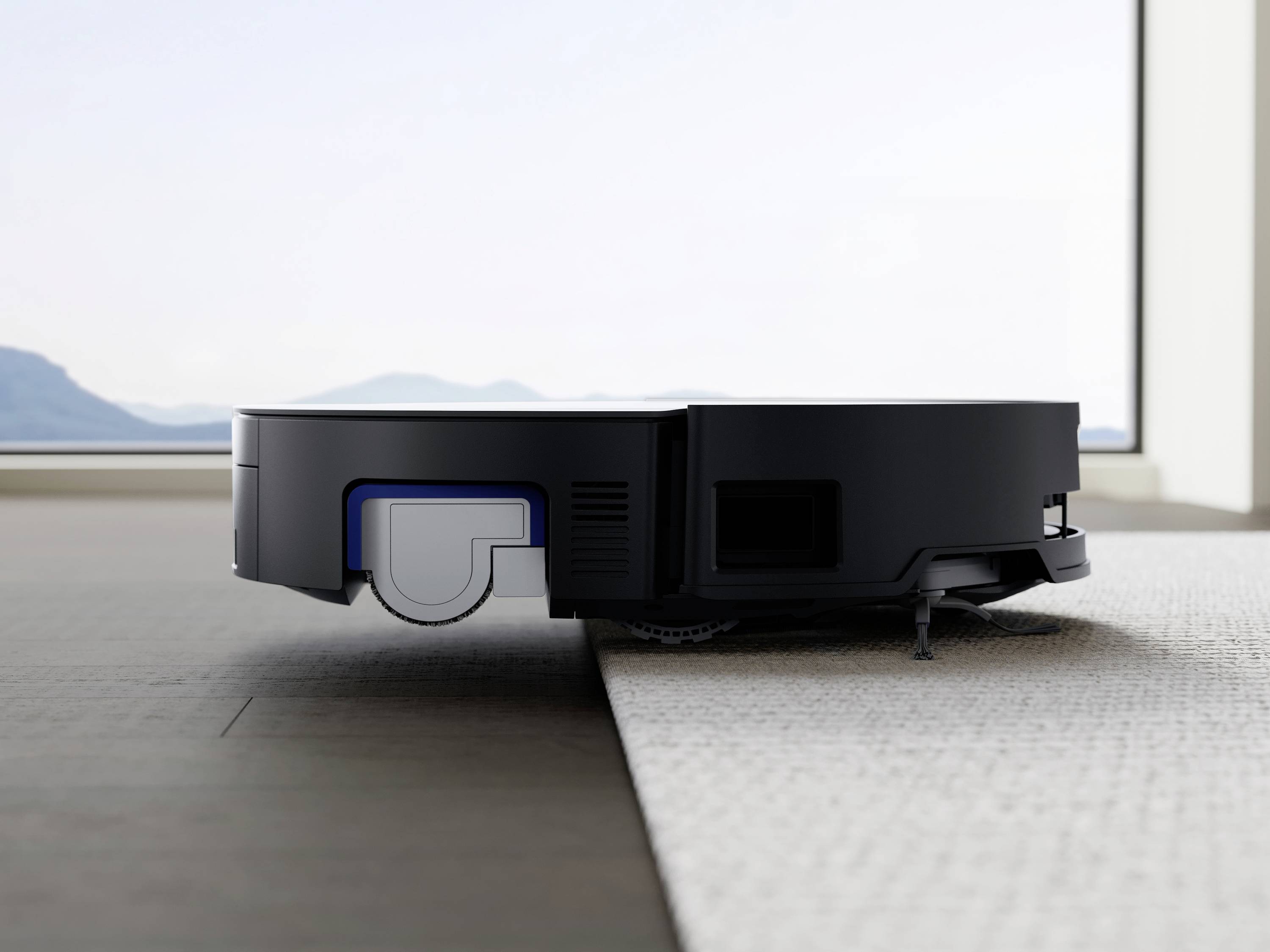 Een robotstofzuiger beweegt zich van een harde houten vloer naar een tapijt in een modern ingerichte ruimte met uitzicht op bergen.