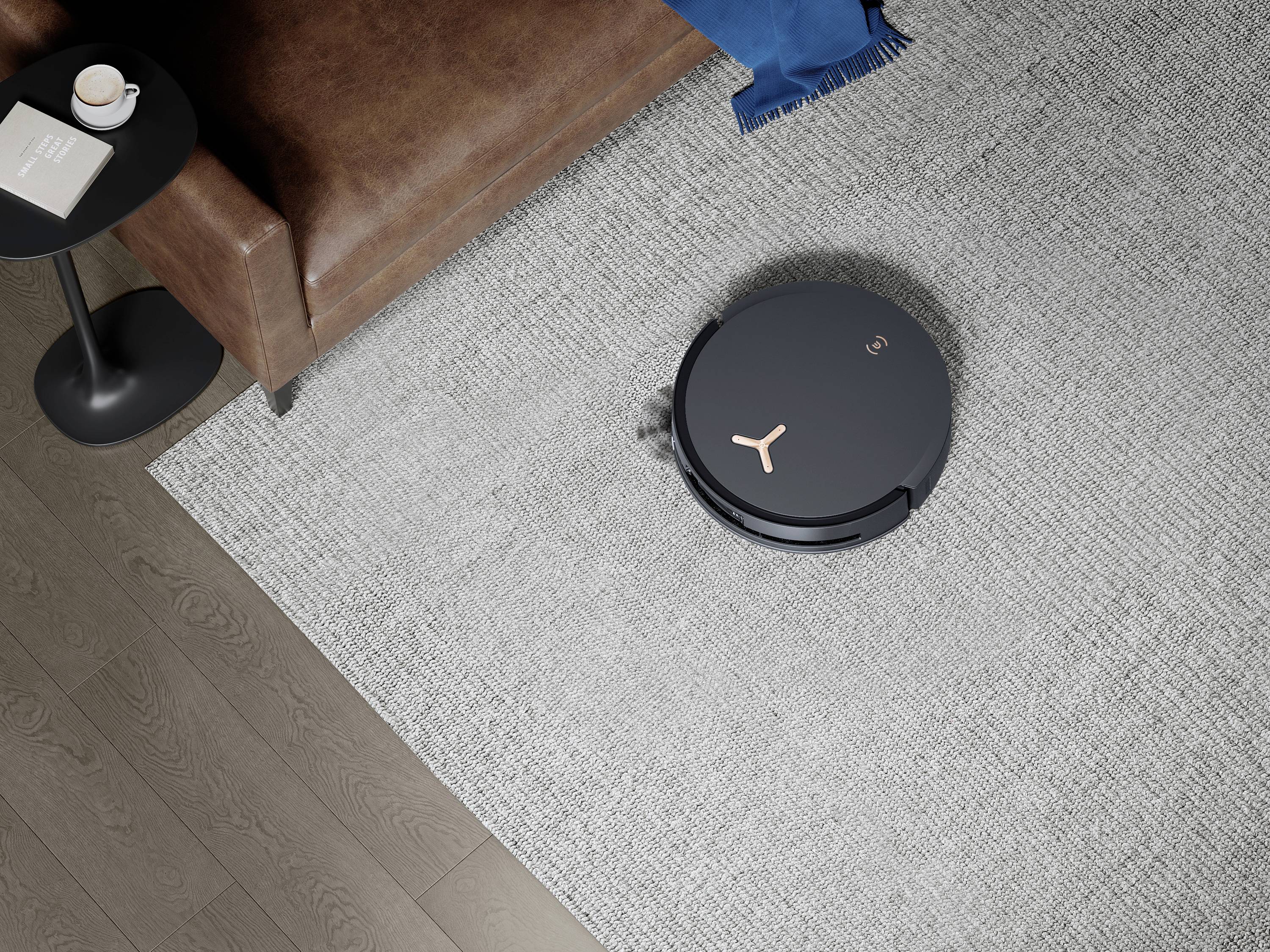 Een ronde robotstofzuiger reinigt een grijze vloerbedekking naast een bruine leren bank. Naast de bank staat een bijzettafel met een boek en een kopje.