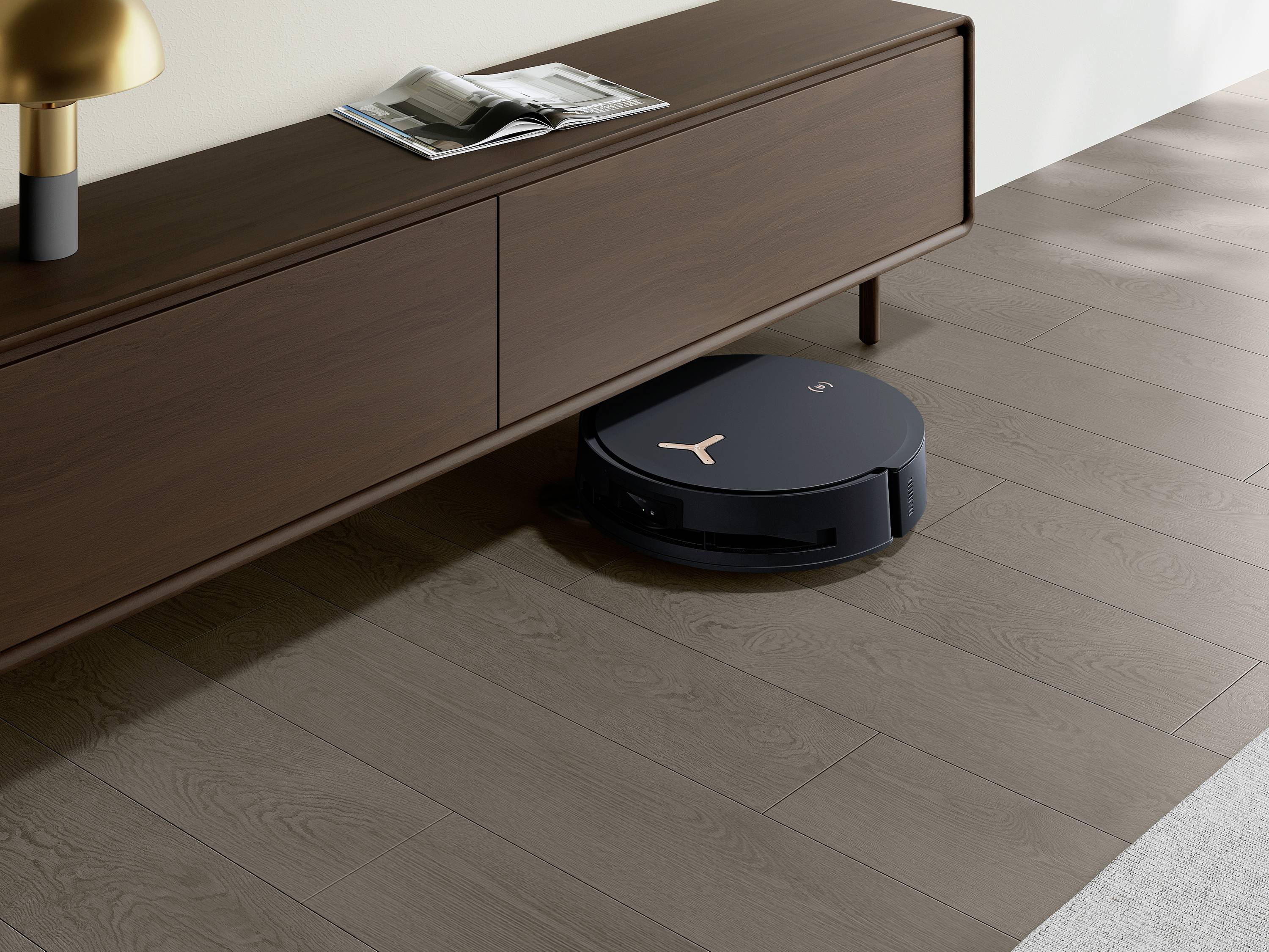 Een zwarte robotstofzuiger reinigt de vloer onder een modern, houten dressoir in een lichte ruimte.