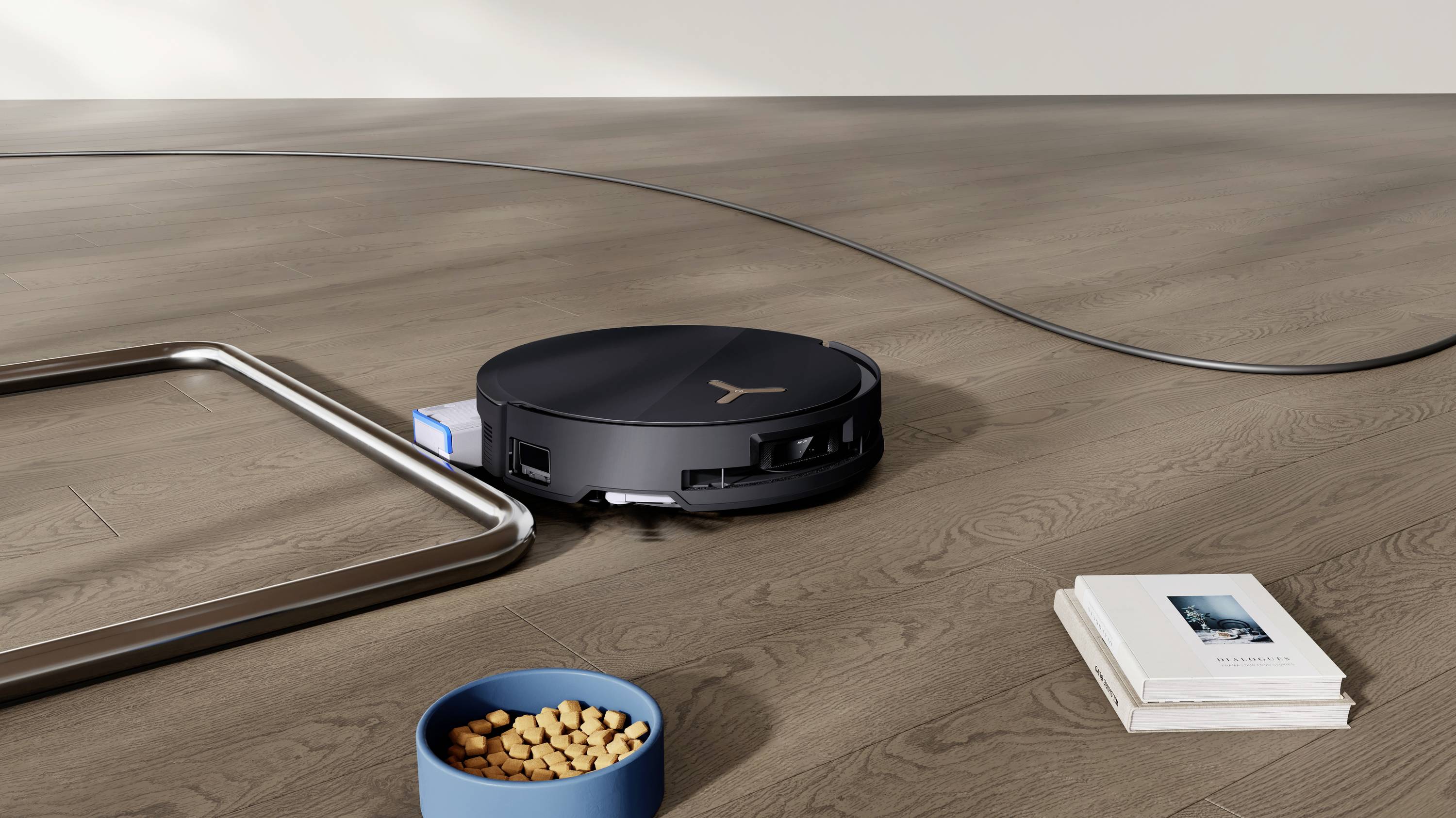 Een robotstofzuiger rijdt over een houten vloer langs een voerbak en boeken terwijl hij stof en vuil opzuigt.