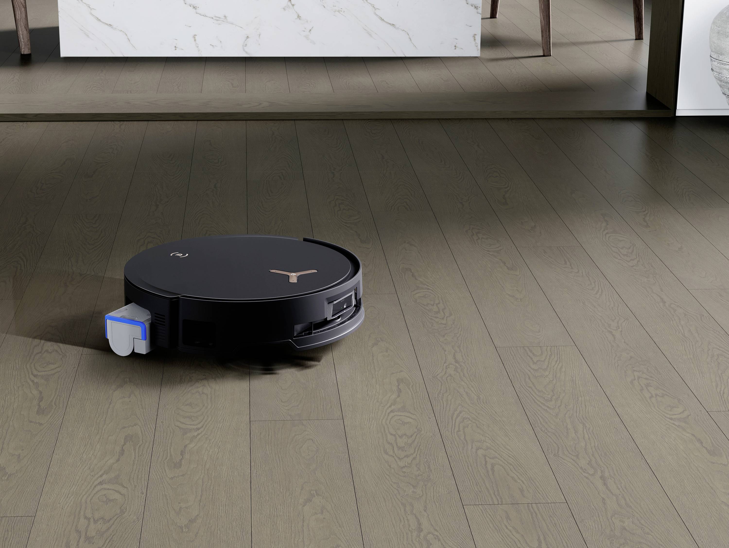 Een ronde robotstofzuiger reinigt een houten vloer in een moderne ruimte. Op de achtergrond zijn tafelpoten en een muur zichtbaar.
