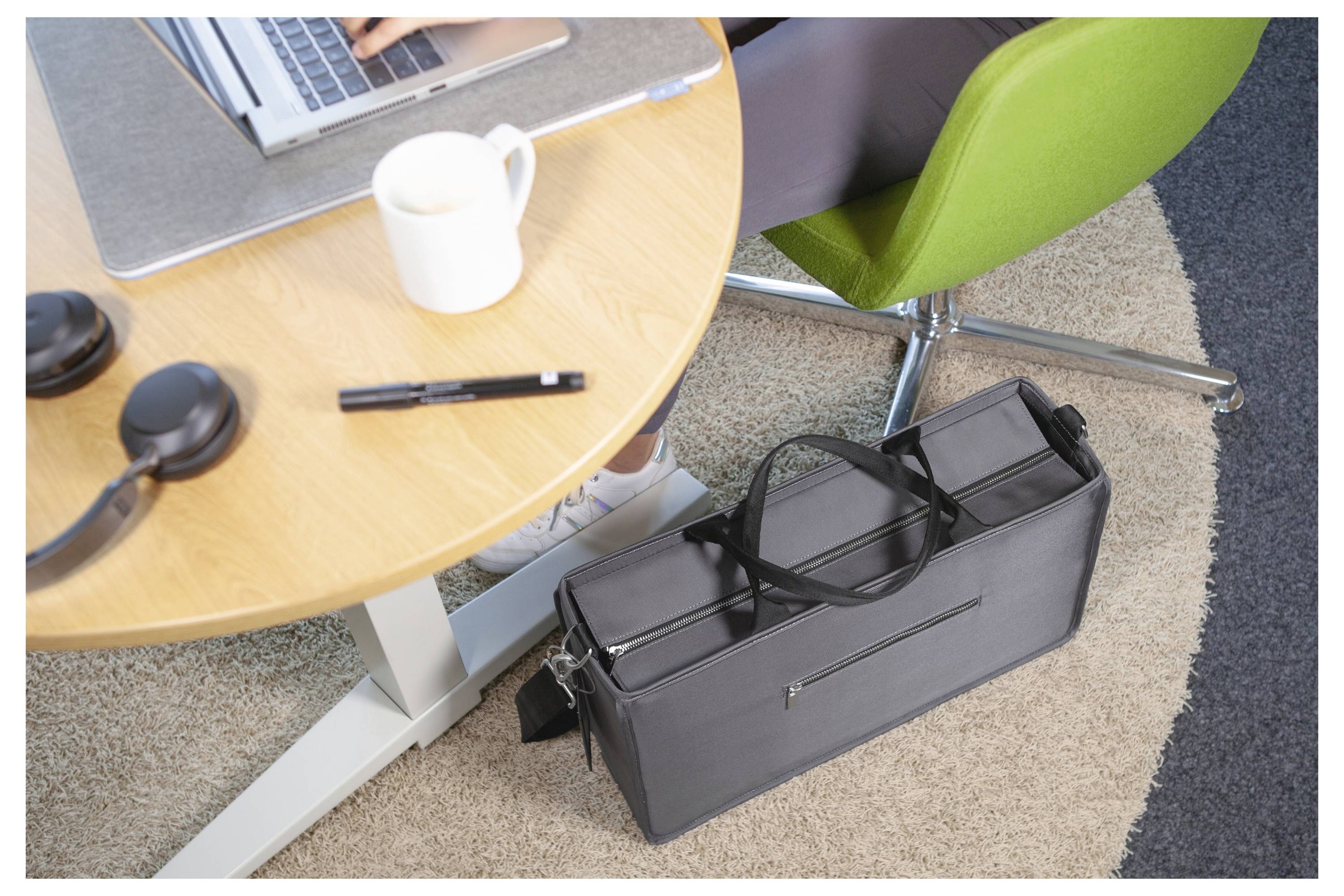 Kantoorvergaderstafel met laptop, koffiekopje, pen, headset en een aktentas op de grond naast een groene stoel.