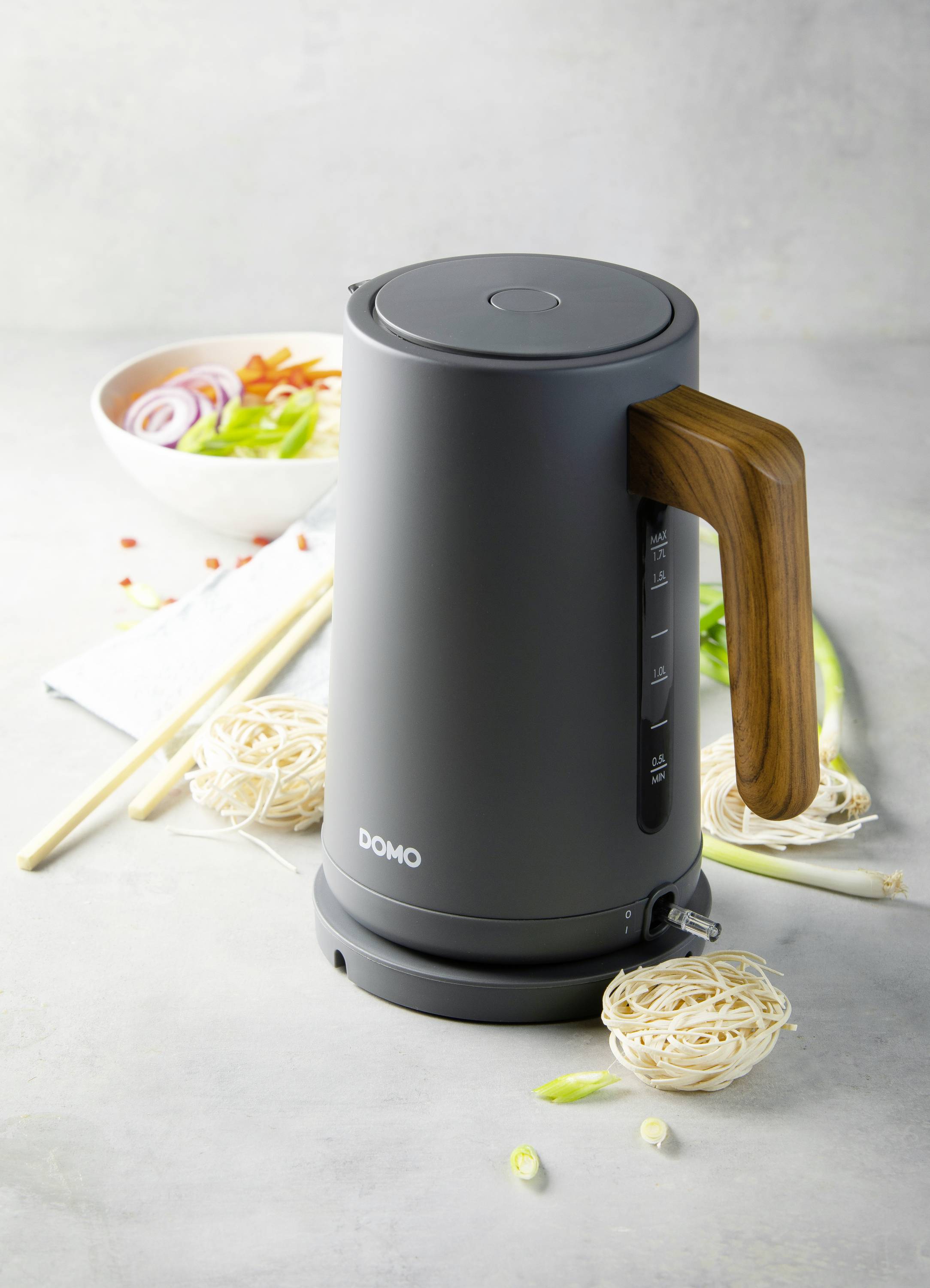 Een zwarte waterkoker met houten handvat staat op een tafel, omringd door pasta en groenten. Op de achtergrond een kom met gesneden groenten.