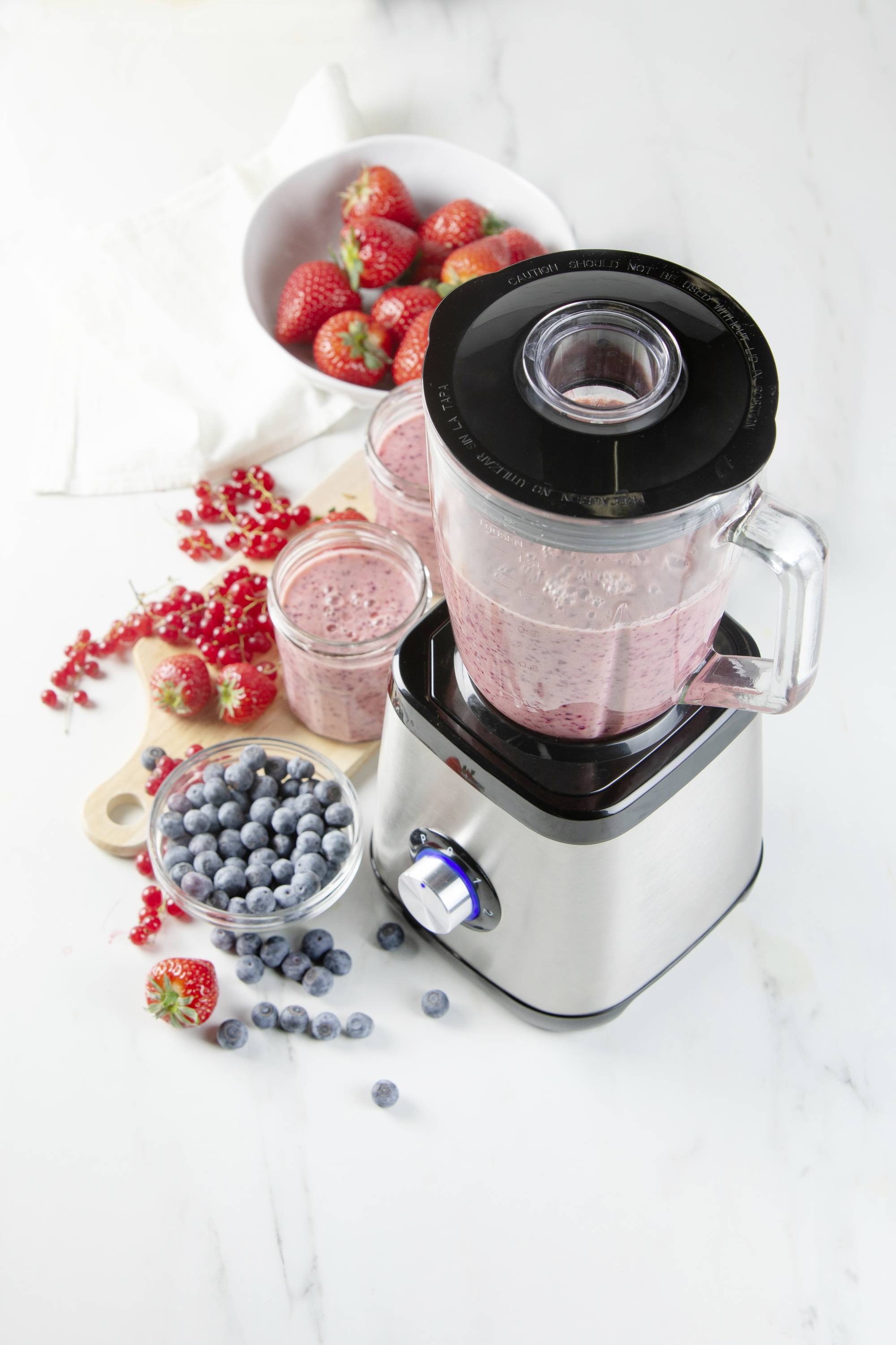 Een blender gevuld met een rode smoothie; ernaast schaaltjes met aardbeien, bosbessen en rode bessen op een witte tafel.