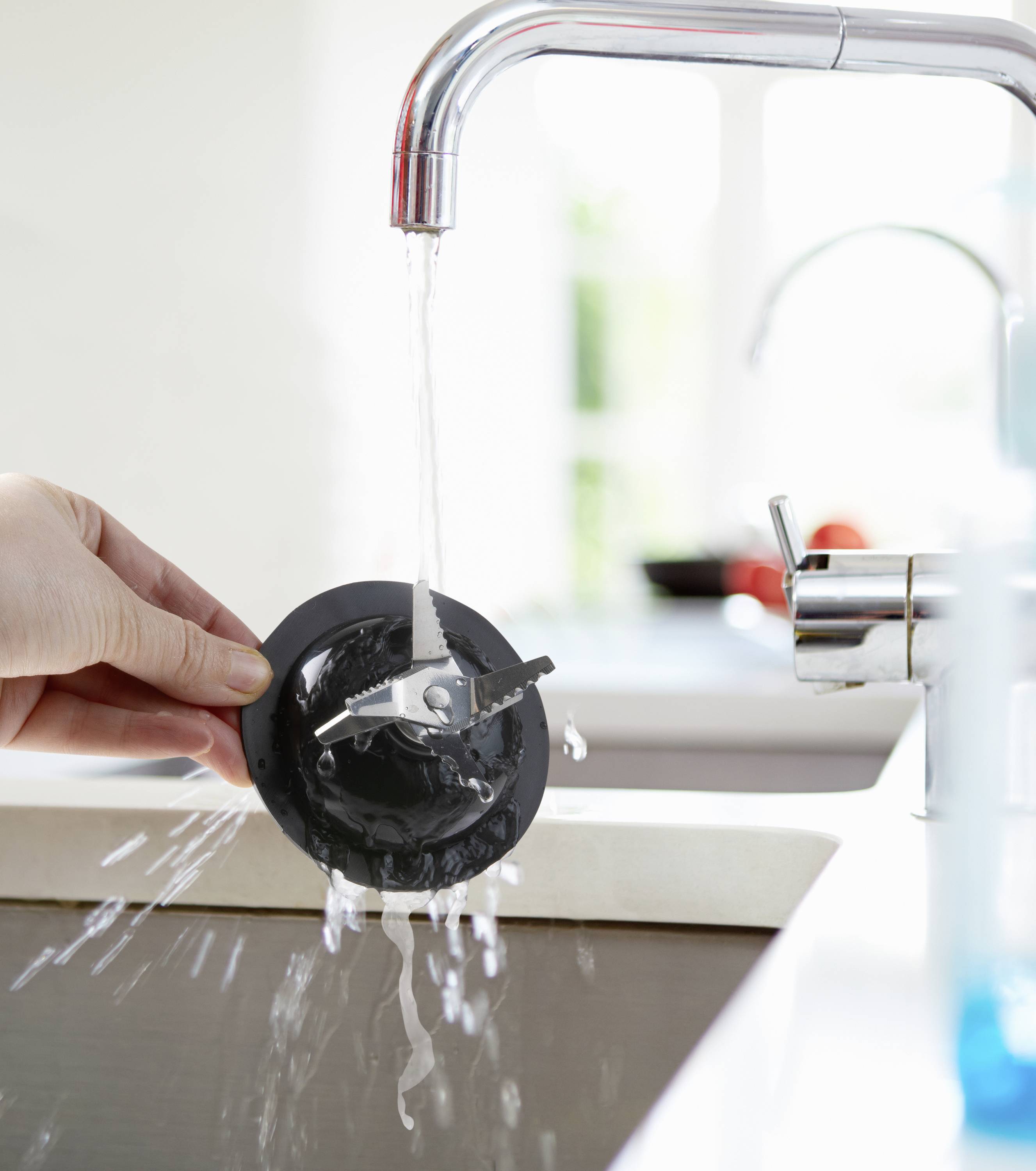 Een hand houdt een afvoerstop onder stromend water om deze schoon te maken. Op de achtergrond is een onscherp raam zichtbaar.