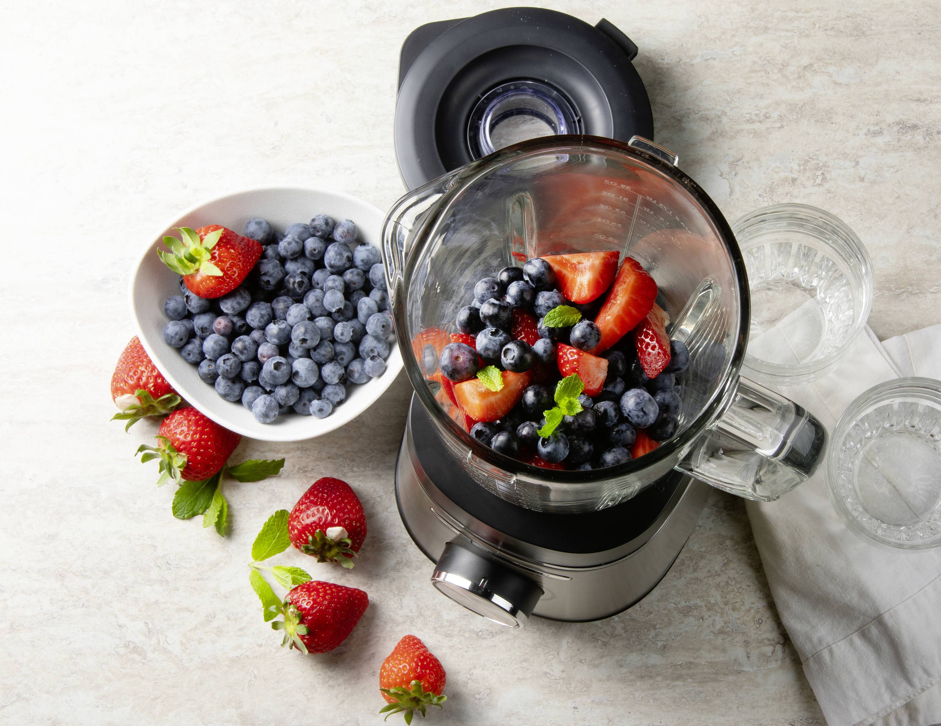Een blender vol aardbeien en bosbessen, naast een kom met extra bessen op een tafel.