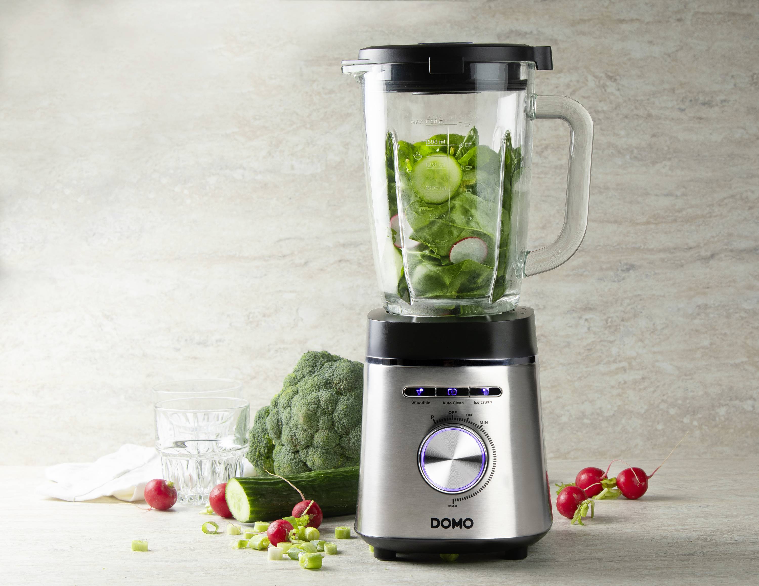Een zilveren blender met verse groenten erin, waaronder komkommerschijfjes en bladeren. Ernaast broccoli, radijsjes en een glas water.