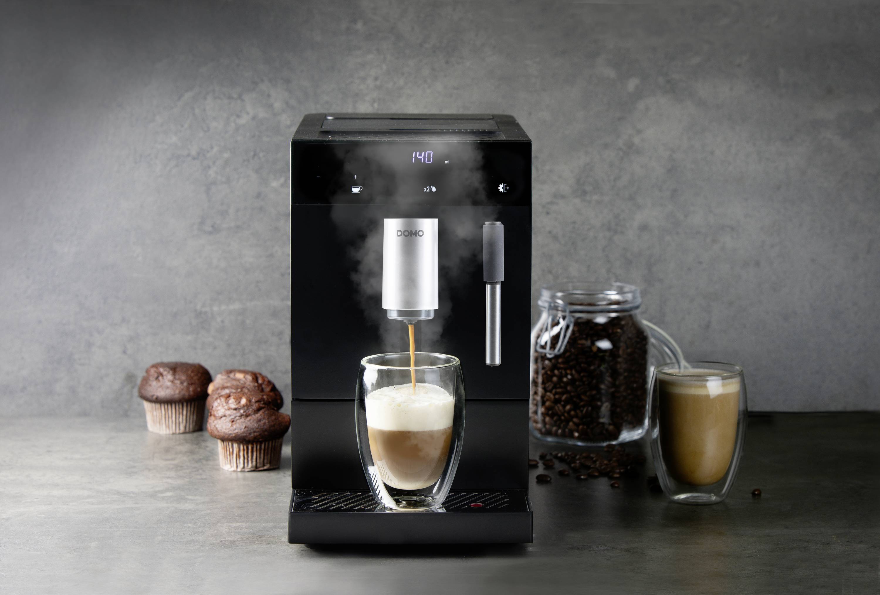 Een zwarte koffiemachine zet koffie in een glas. Ernaast staan twee muffins, een glas met koffiebonen en nog een glas koffie.