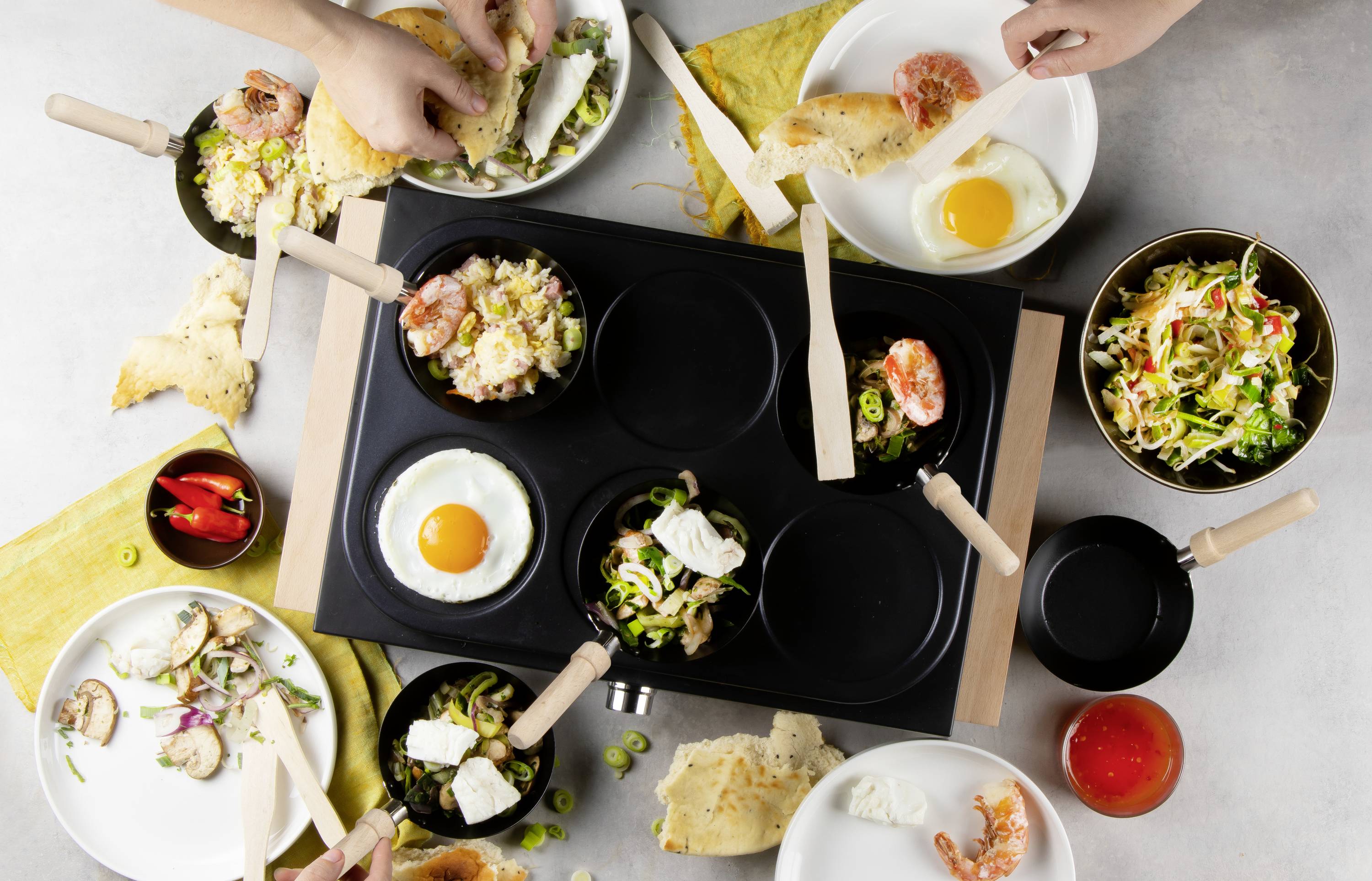 Een tafel met serveerplannen, spiegeleieren, salade, broodstukken en groenten. Handen grijpen naar het eten. Scenario voor het delen van een maaltijd.