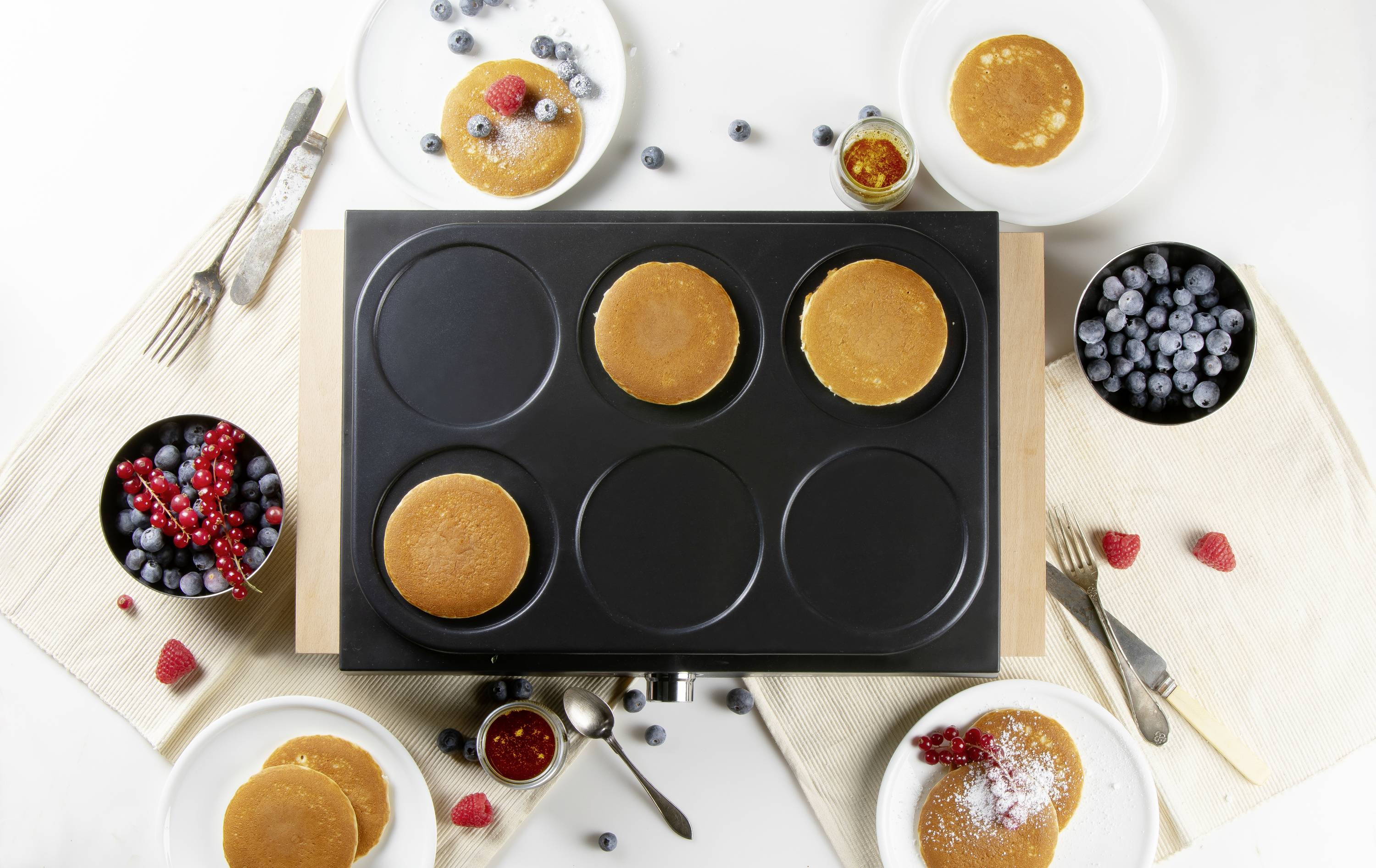 Een pan met meerdere ronde pannenkoeken, omringd door borden met bessen en bestek op een tafel.