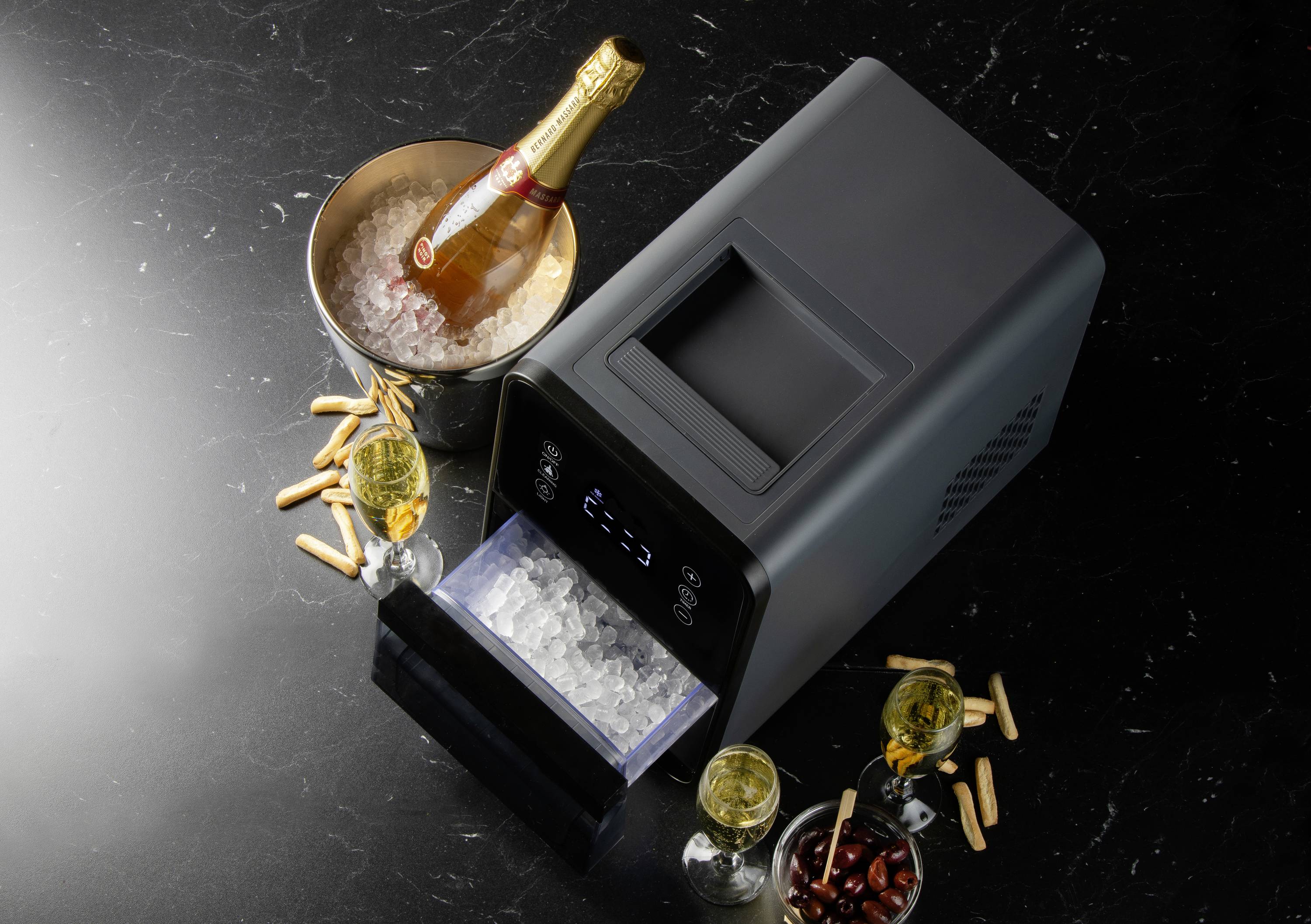 IJsmachine op een zwarte ondergrond, met drie gevulde champagneglazen, een champagnefles in een ijsemmer en enkele hapjes.